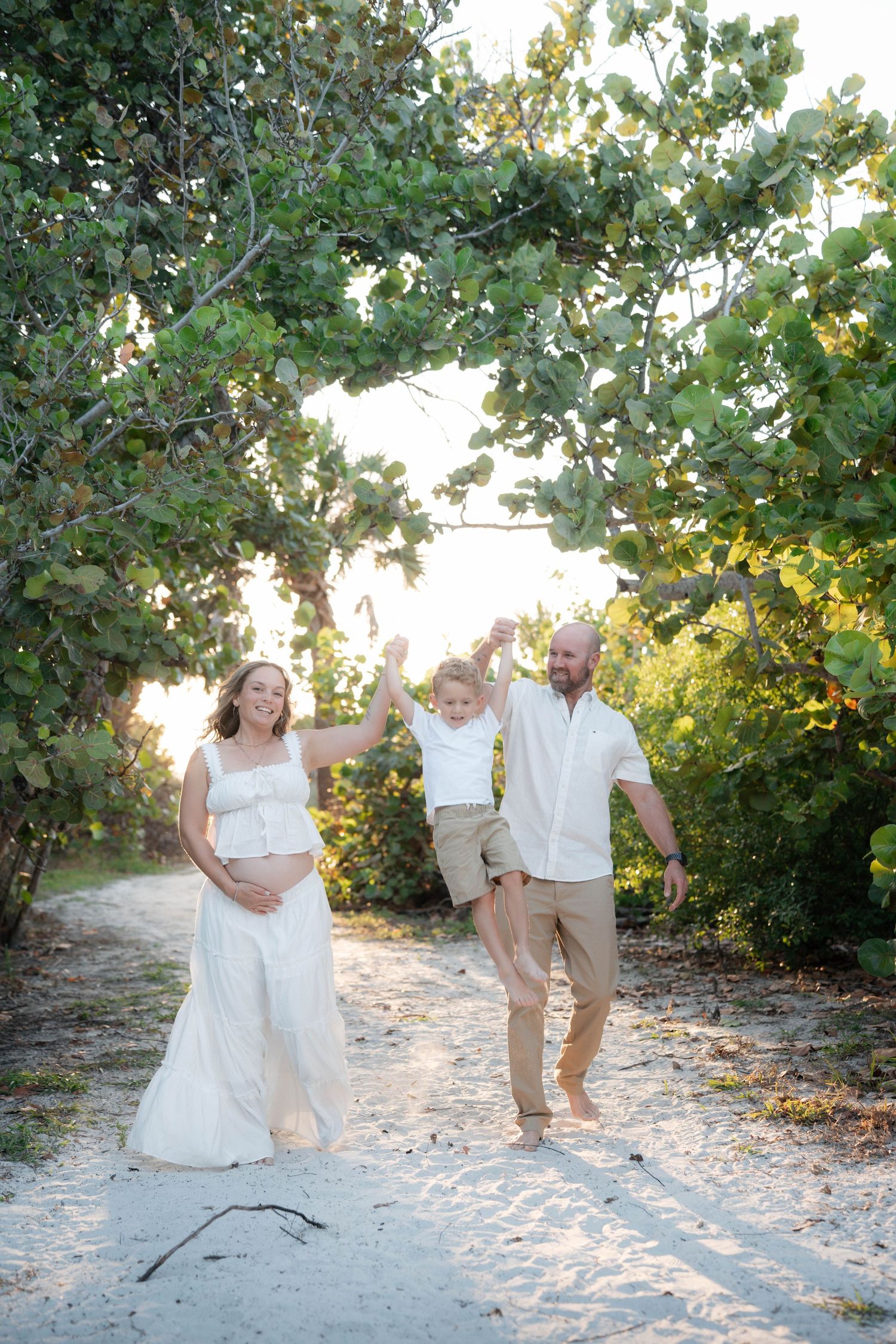 Pregnant woman in white dress and bald man swing toddler on sandy beach path surrounded by lush green trees.
