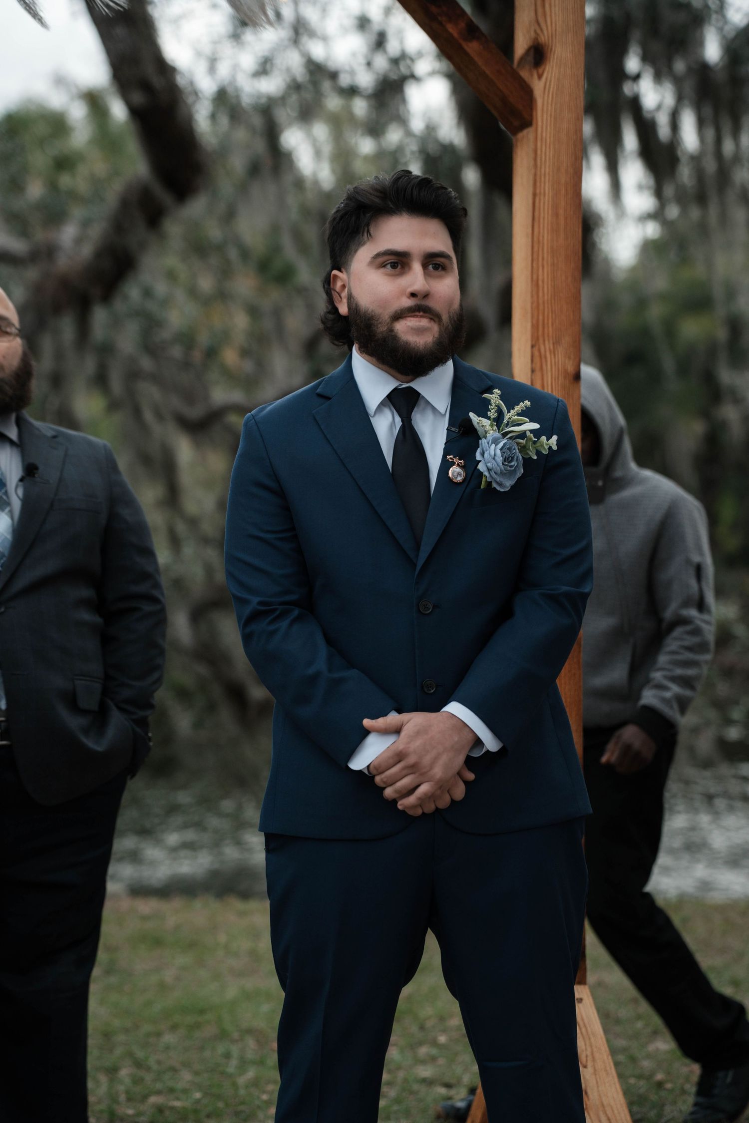 Groom in navy suit with floral boutonniere stands at outdoor wedding altar among Spanish moss trees.