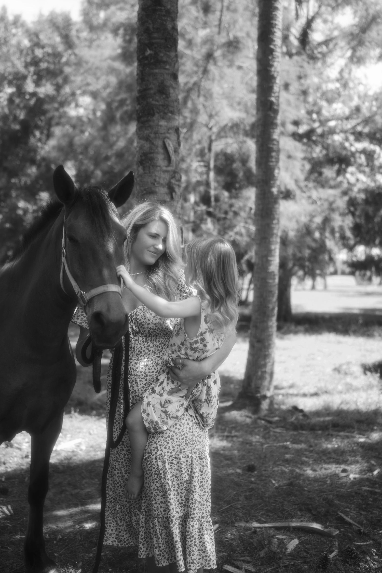 Black and white photo of a woman holding a child beside a horse among tall trees in a serene outdoor scene.
