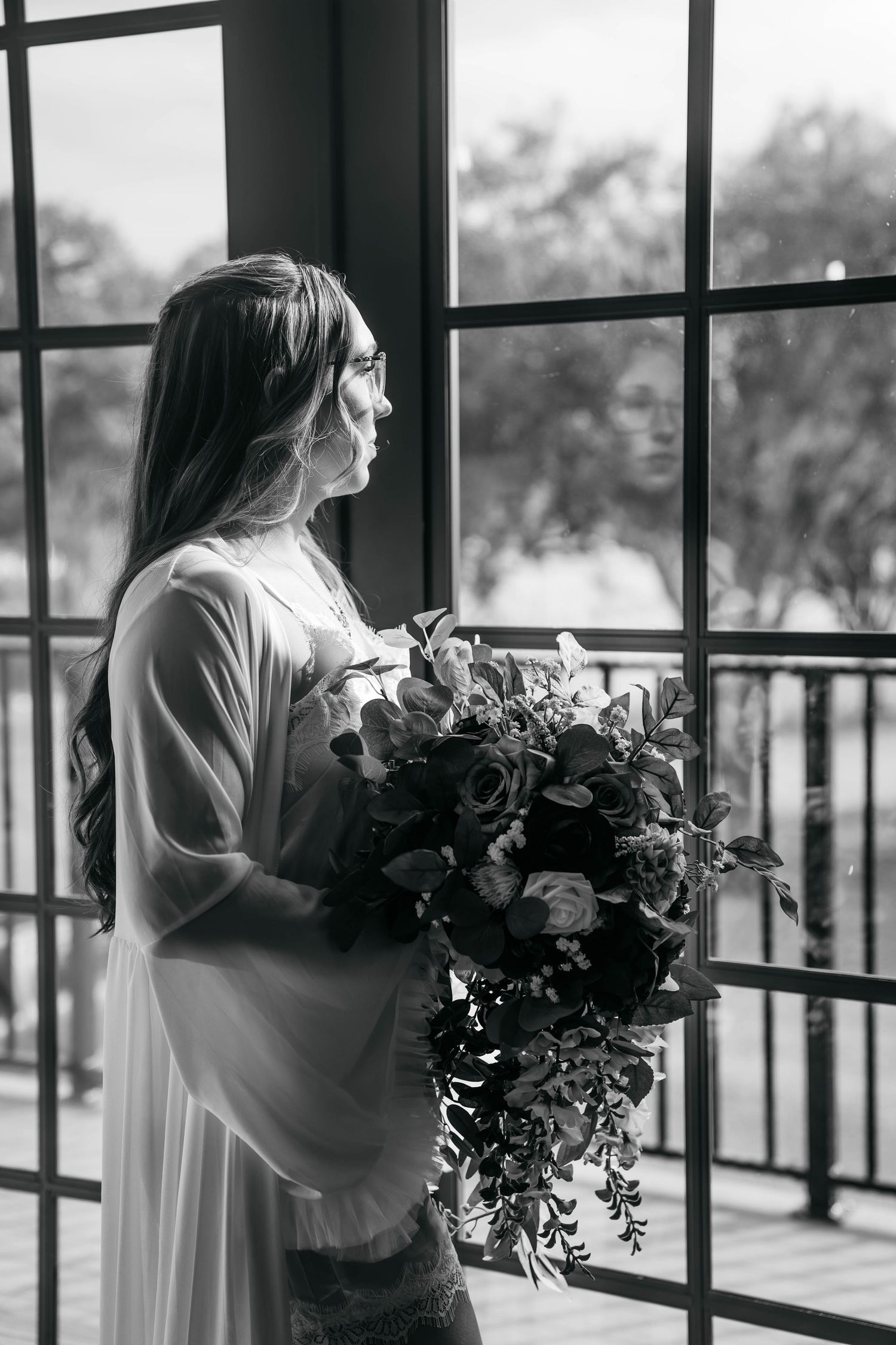 Black and white portrait of bride in robe holding lush cascading bouquet while gazing out large window.