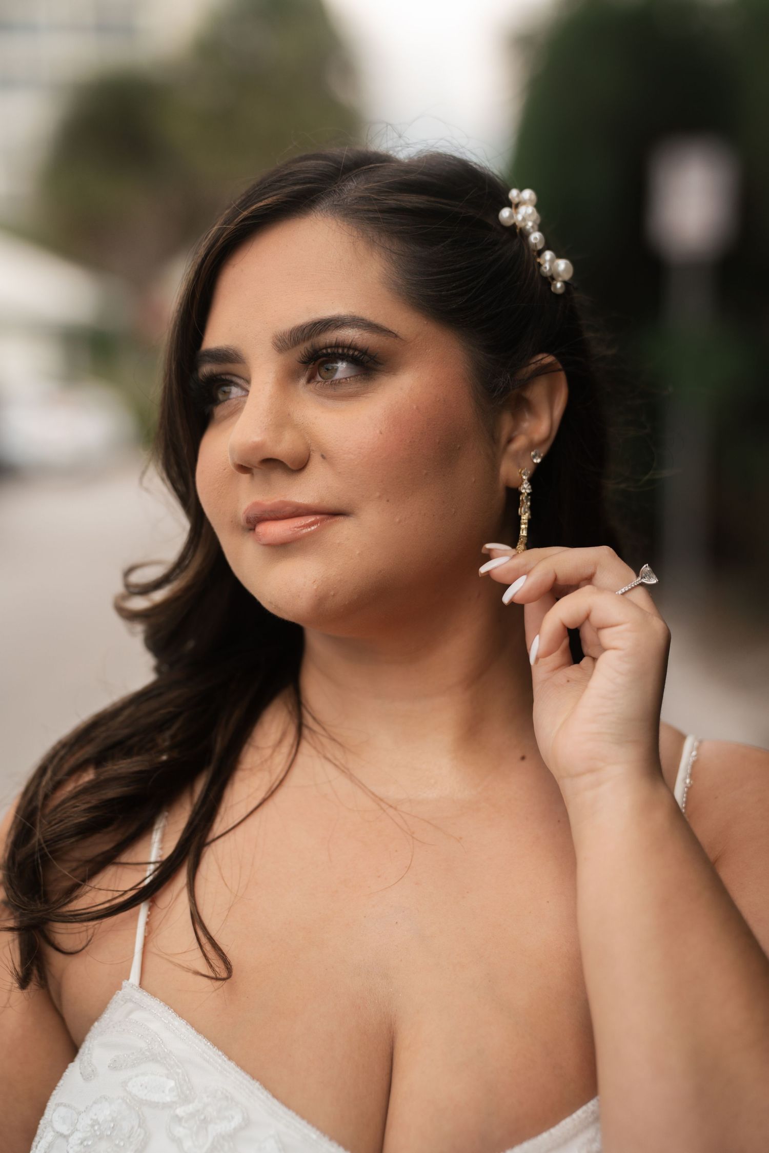 Elegant bride with pearl hair accessories and dangling earrings in white gown poses outdoors.