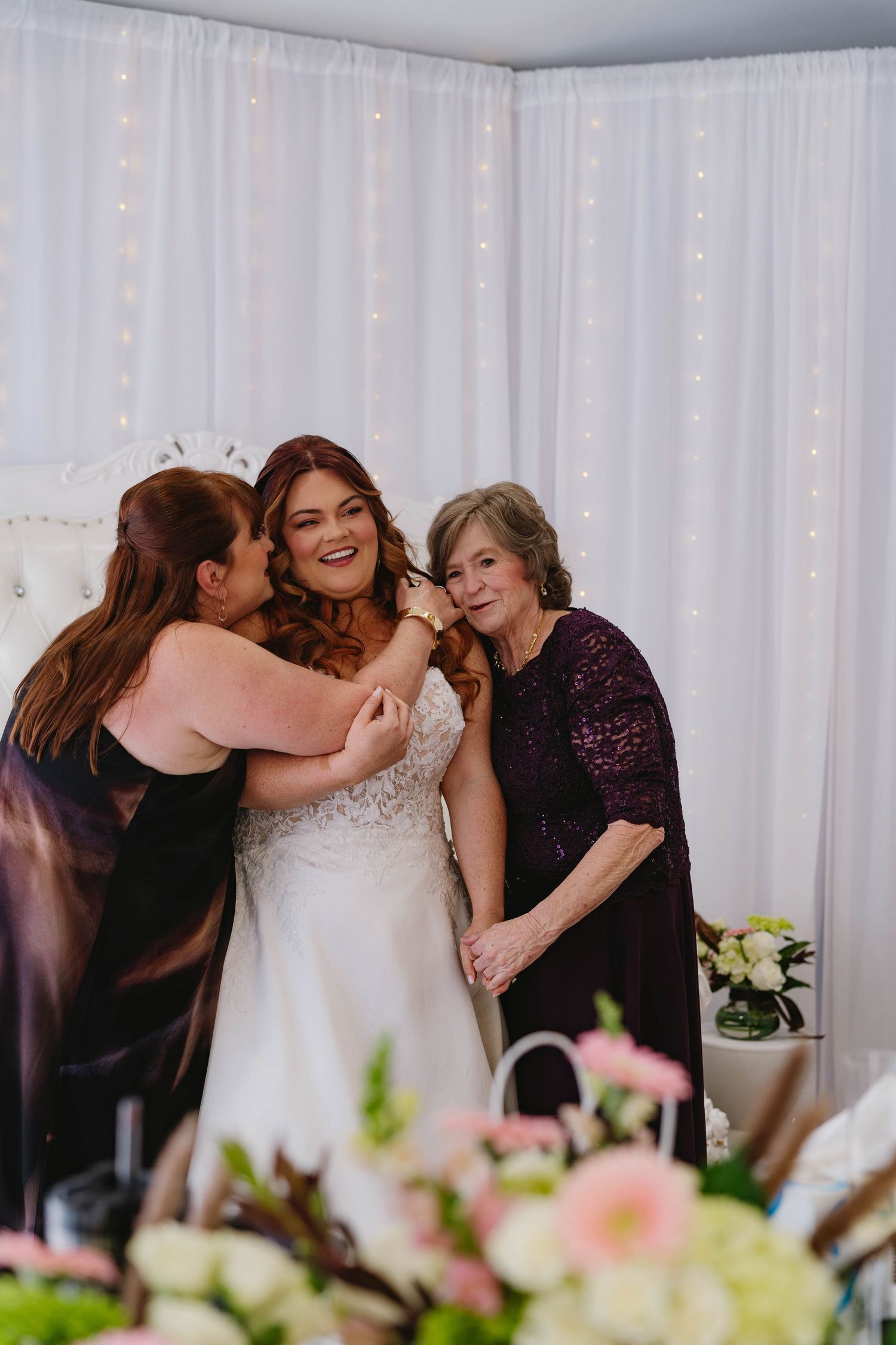 Bride smiling joyfully as two women help her into her lace wedding gown in a fairy-lit bridal suite.