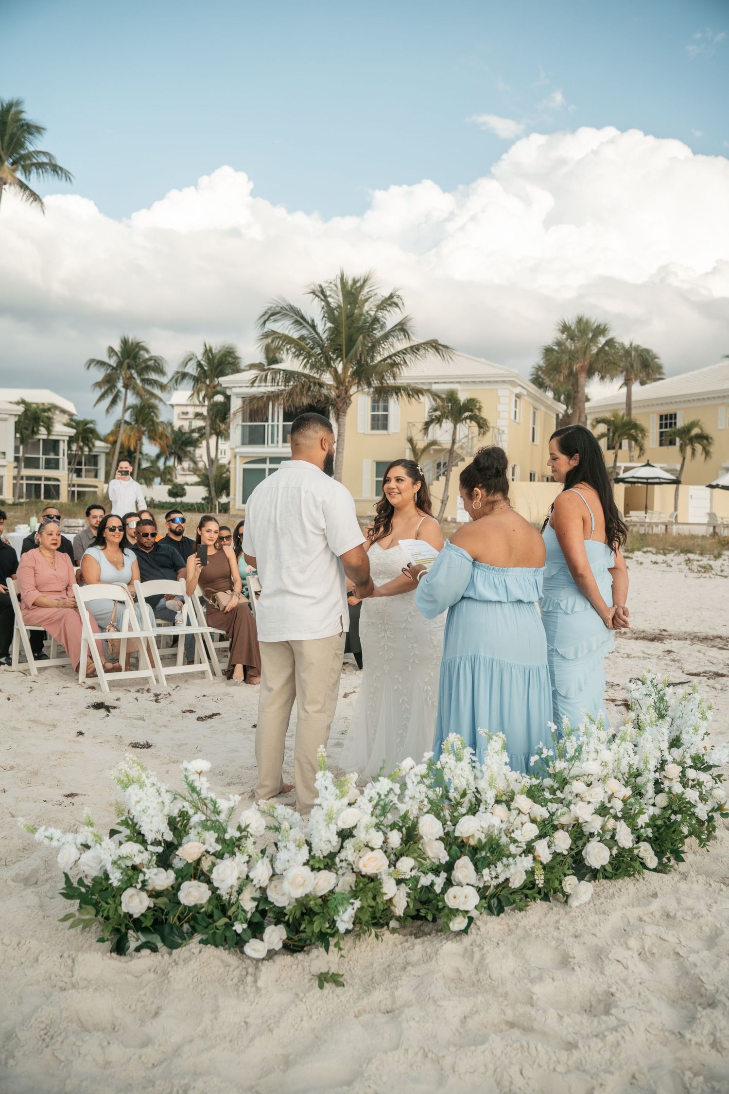 Beach wedding ceremony with couple exchanging vows surrounded by white floral arrangement.