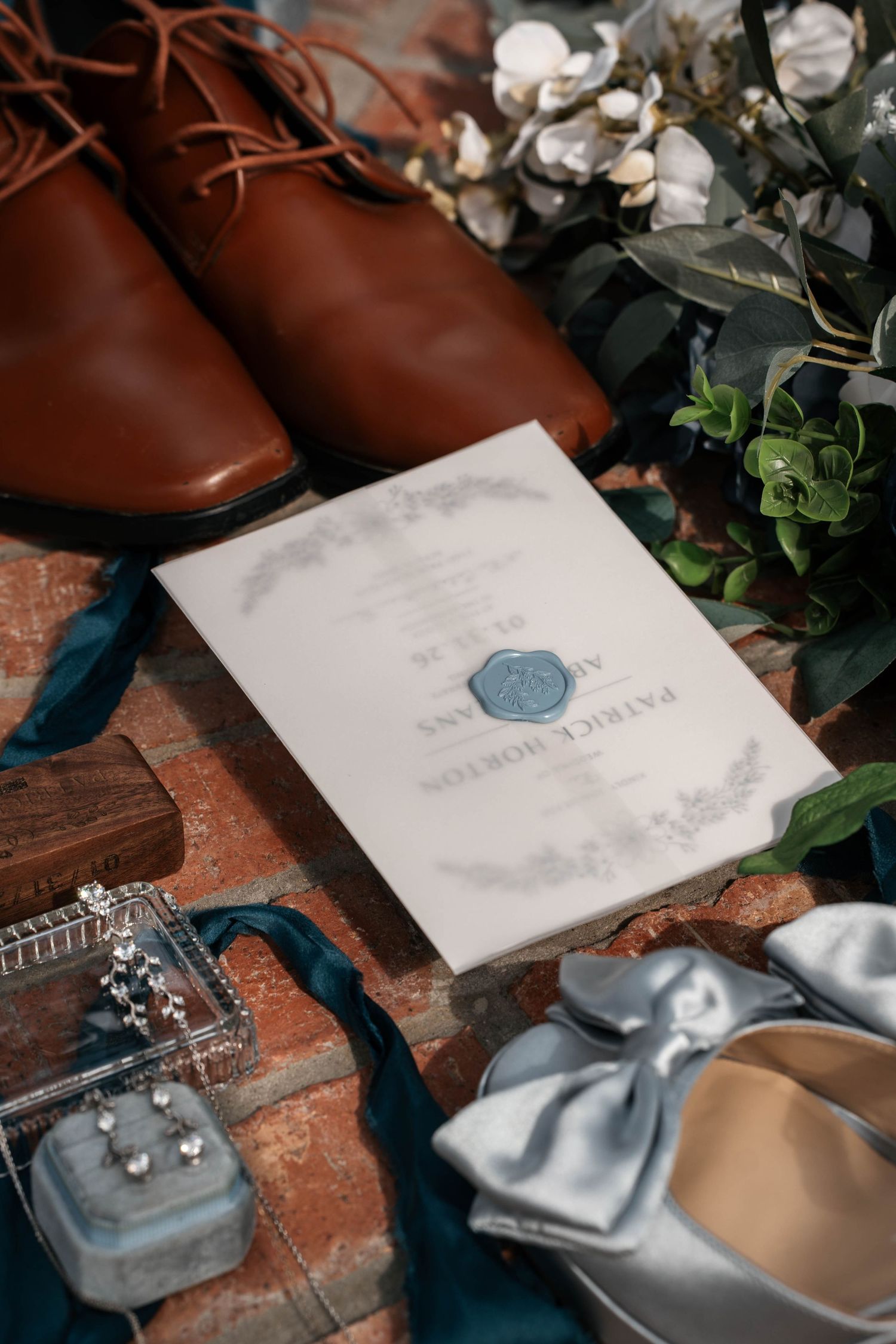 Wedding flat lay with brown leather shoes, invitation, rings, earrings, and blue heels.