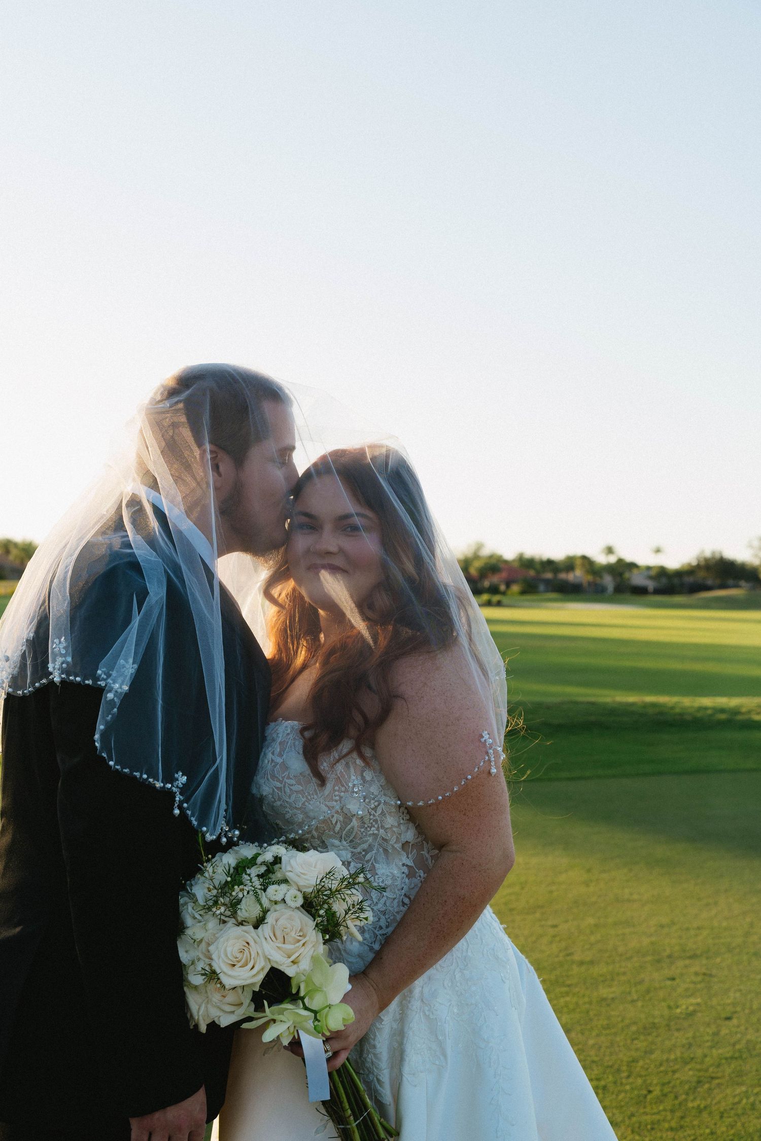Groom kisses bride on cheek under veil as she smiles on sunny golf course.