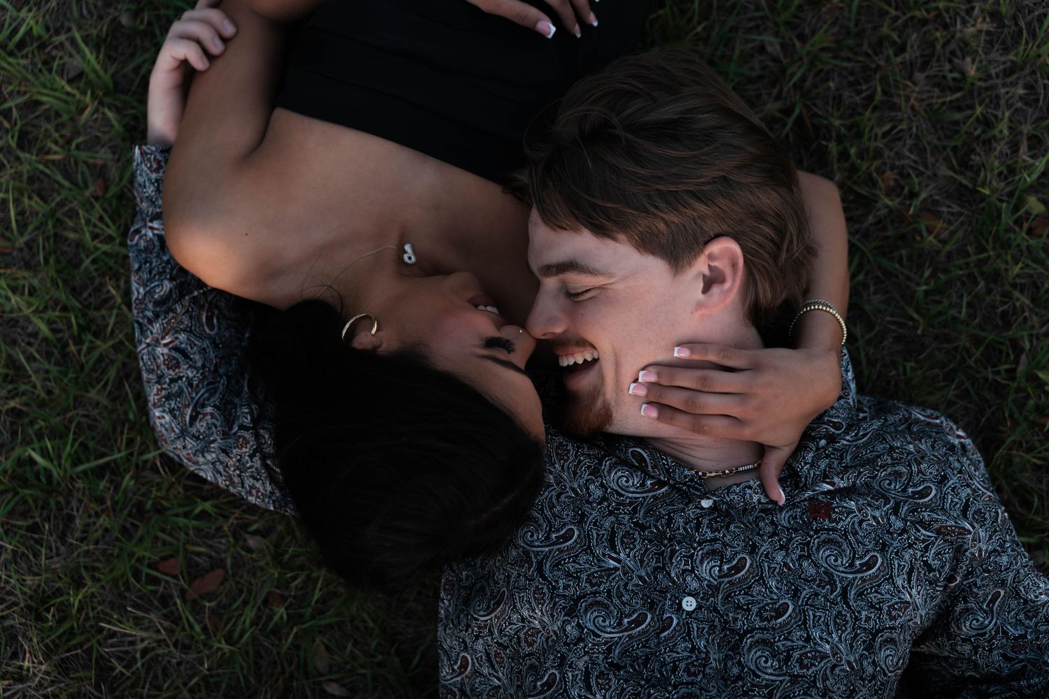 Couple laughs together lying on grass in joyful outdoor engagement photo session.
