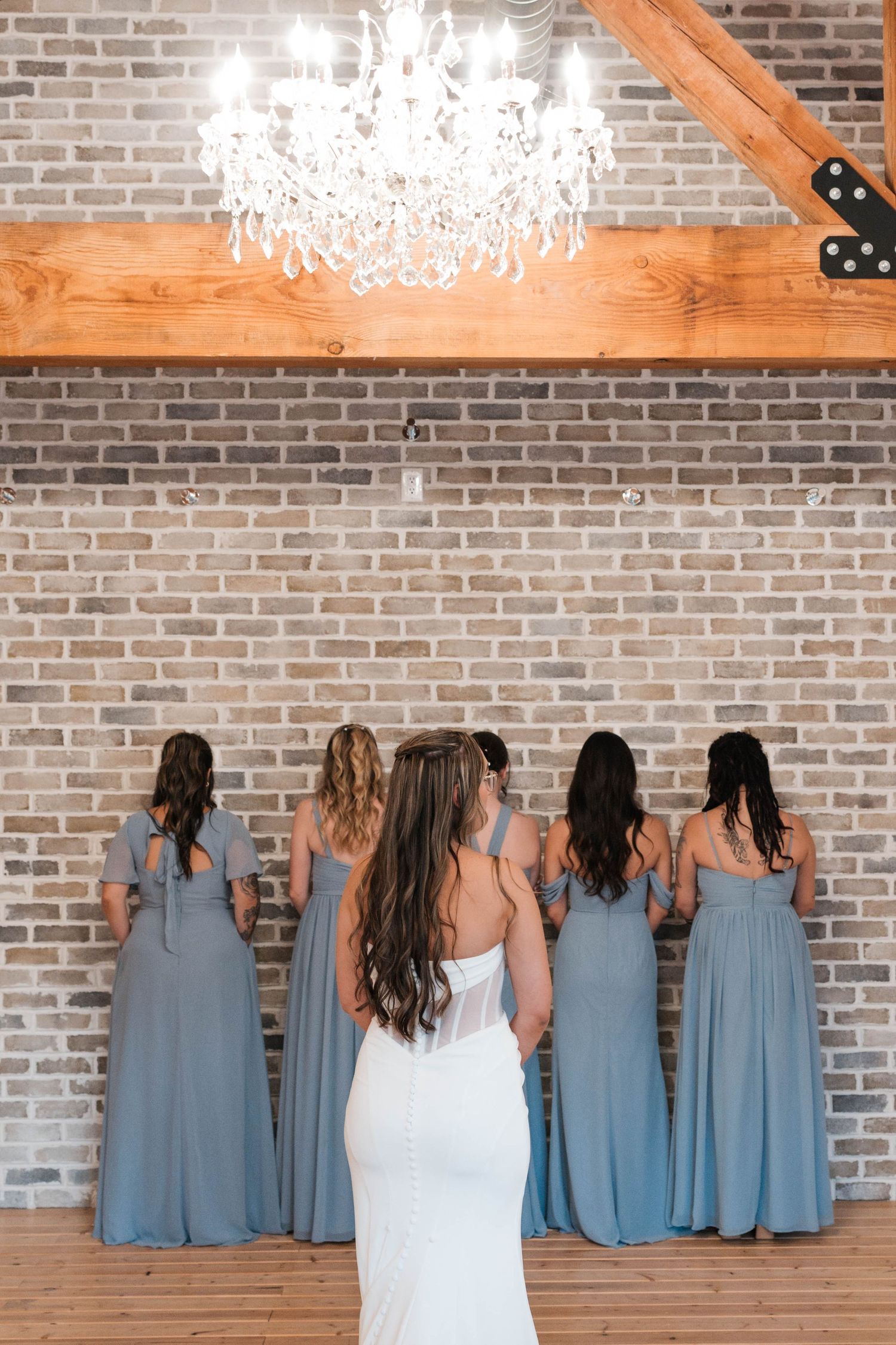Bride stands with four bridesmaids in dusty blue gowns facing brick wall under chandelier.
