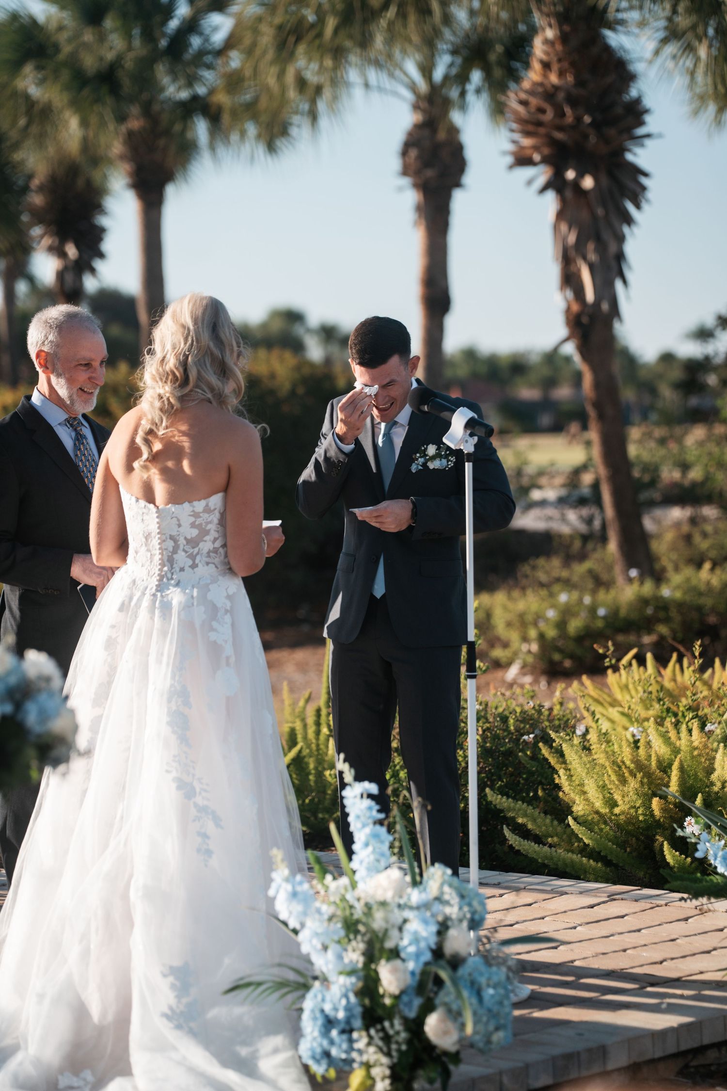 Groom reads emotional vows to bride at outdoor wedding ceremony with palm trees and blue floral arrangements.
