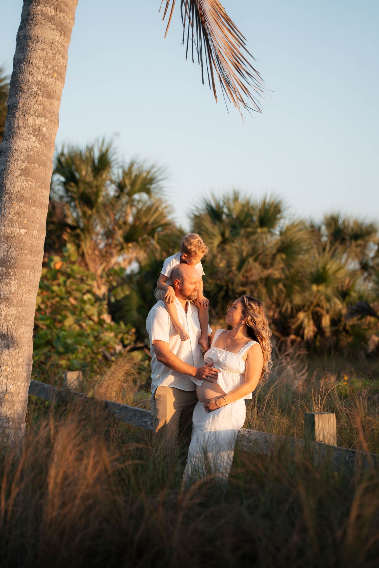 A pregnant woman in a white dress stands with her husband and child on shoulders near palm trees at golden hour.
