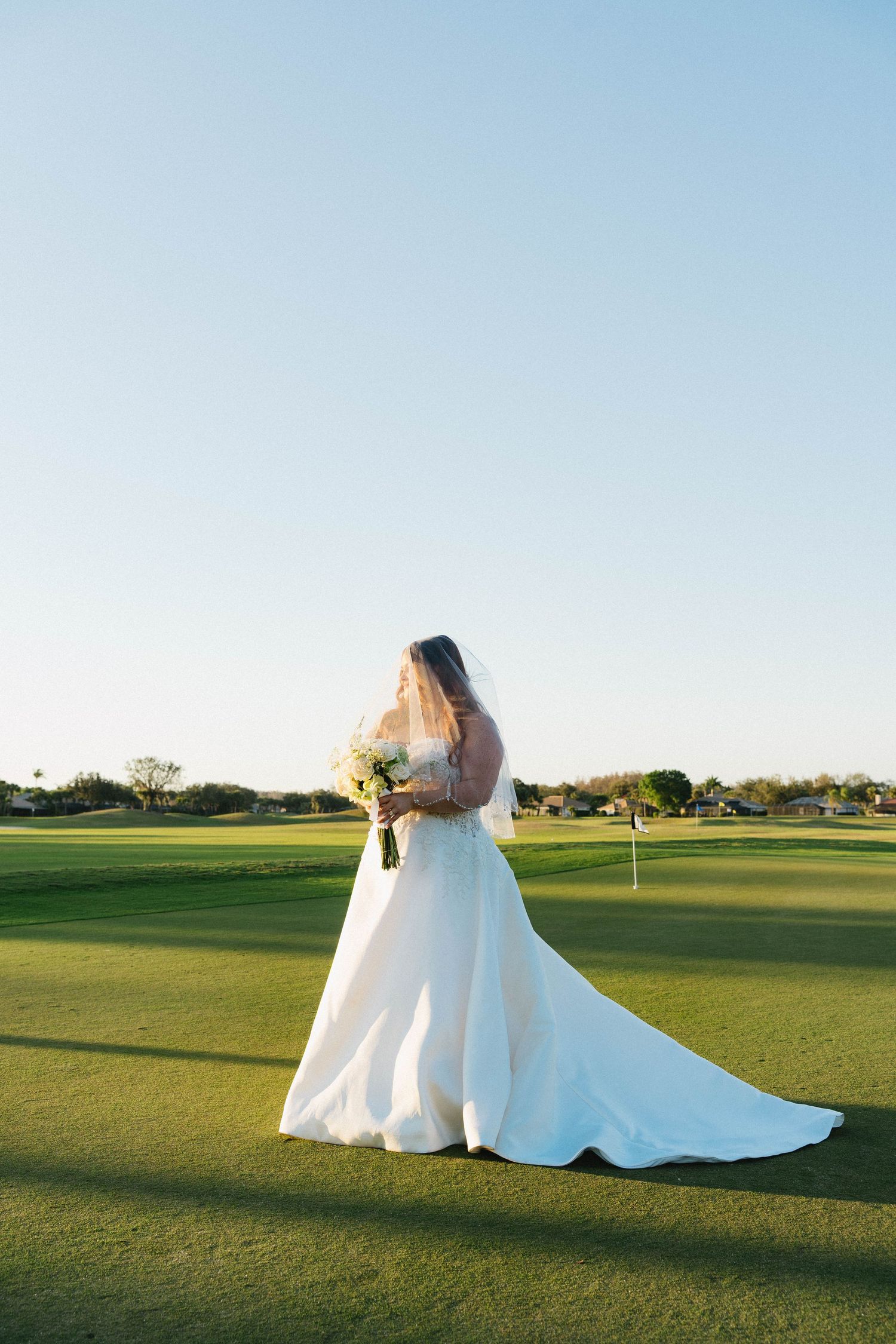 Bride in white gown and veil stands on golf course green holding bouquet at sunset.