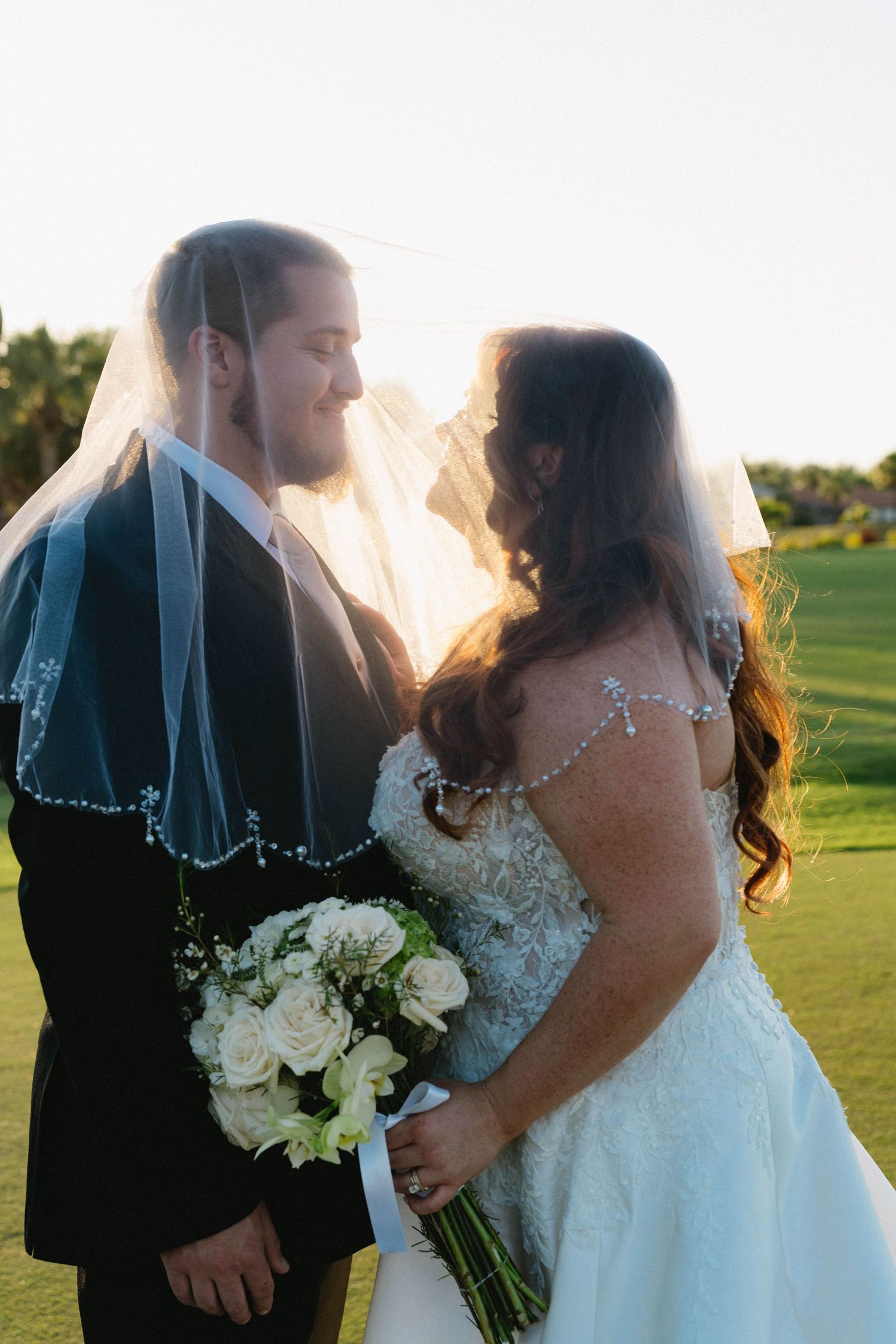 Bride and groom share romantic moment under flowing veil holding white rose bouquet.