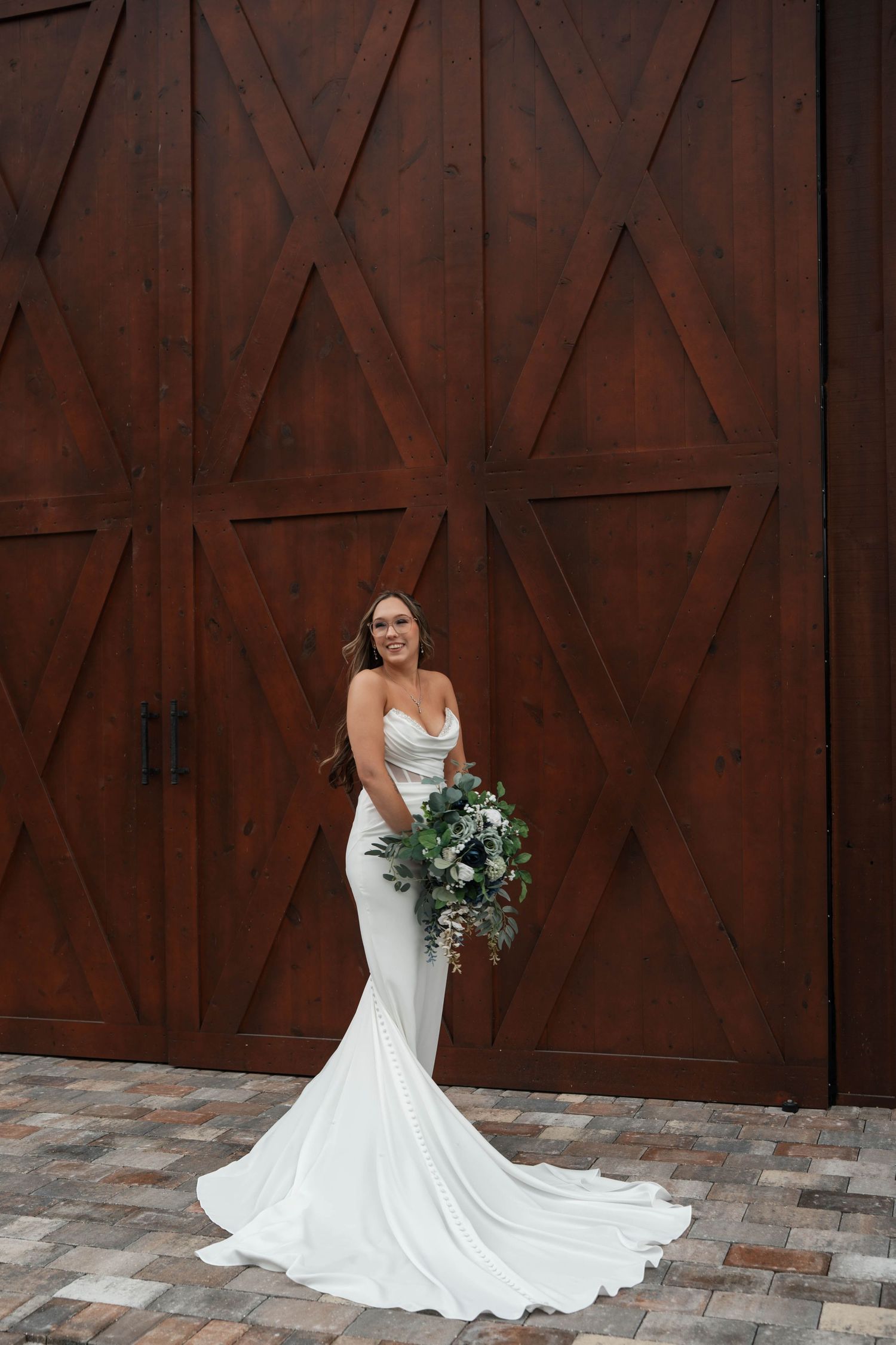 Smiling bride in fitted white gown with long train holds greenery bouquet before rustic barn wood doors.