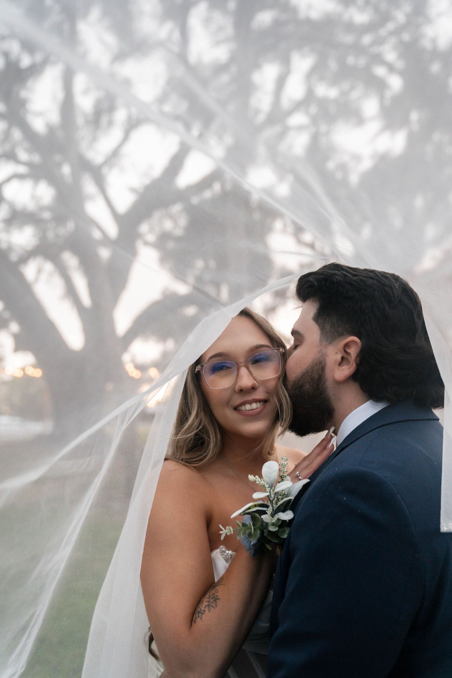 Groom kisses smiling bride on cheek under flowing veil with dramatic tree backdrop at sunset.
