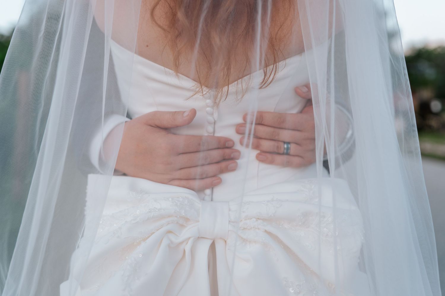 Close-up of bride in white gown with groom's hands around her waist showing wedding ring.