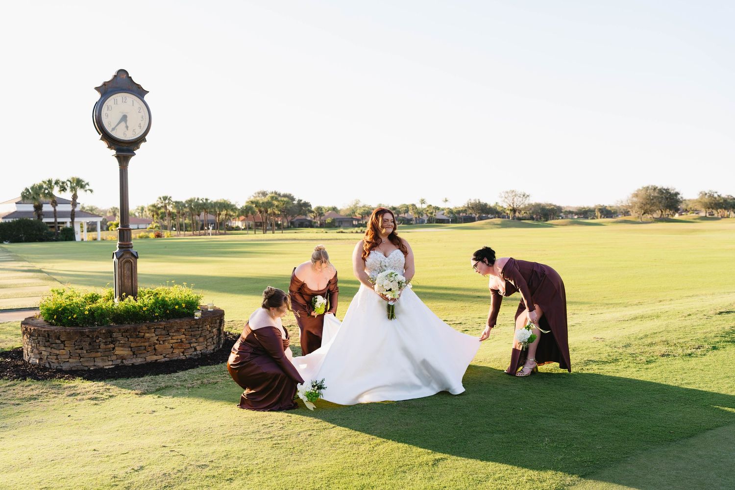 Bridesmaids in burgundy gowns fluffing bride's white dress on a golf course green.