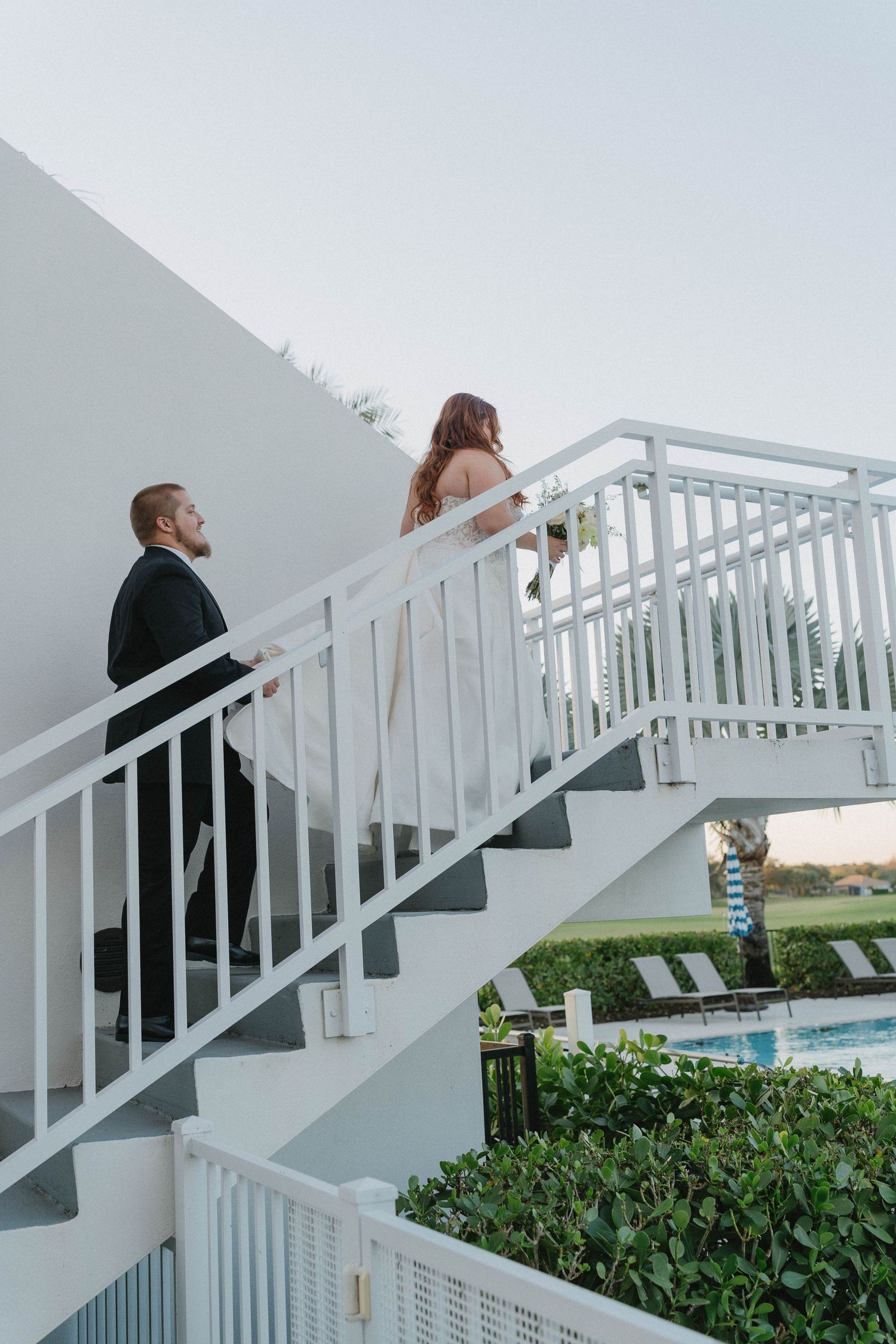 Bride and groom walk up white outdoor staircase near pool at elegant wedding venue.