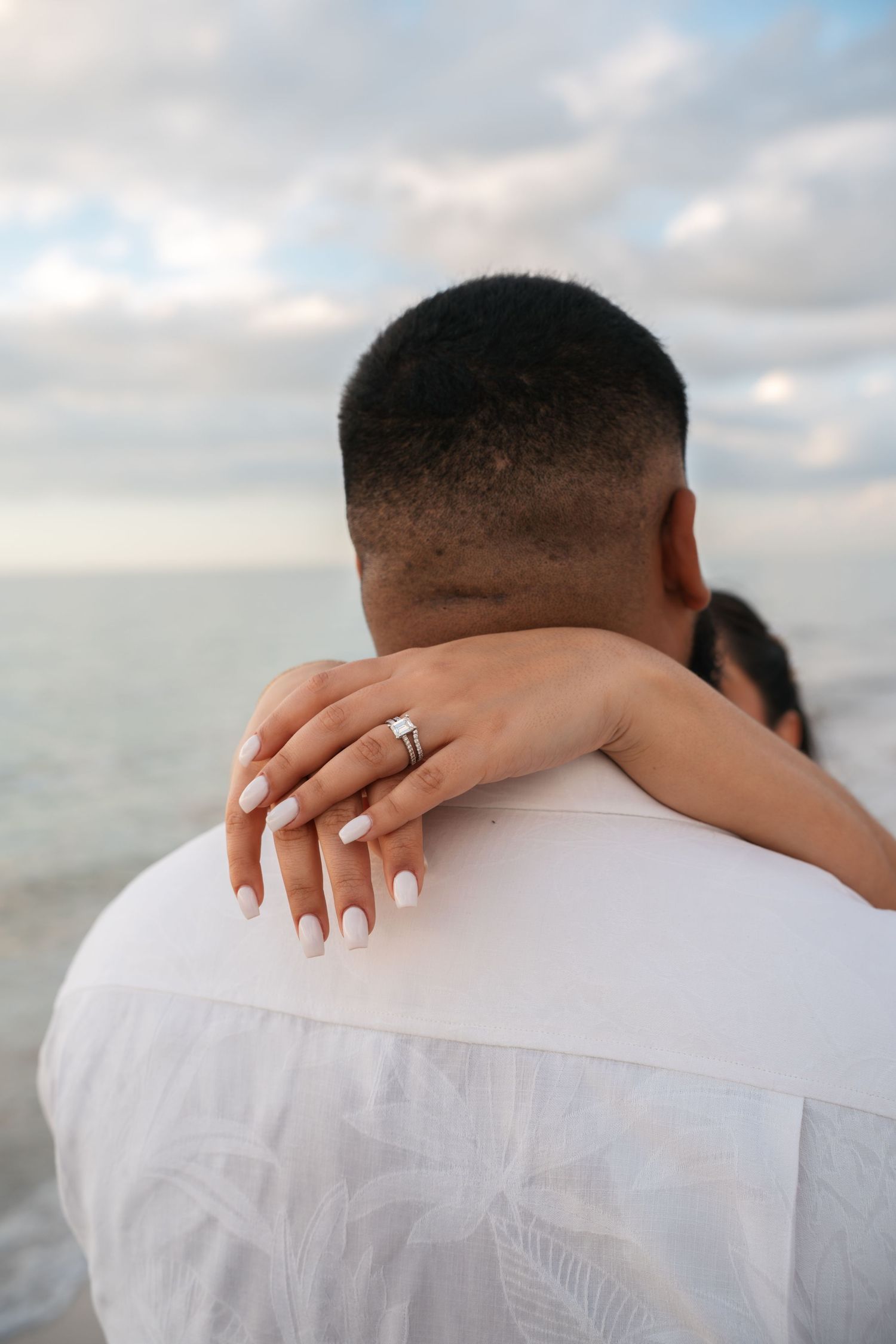 Bride's hand with diamond ring draped over groom's shoulder at beach wedding.