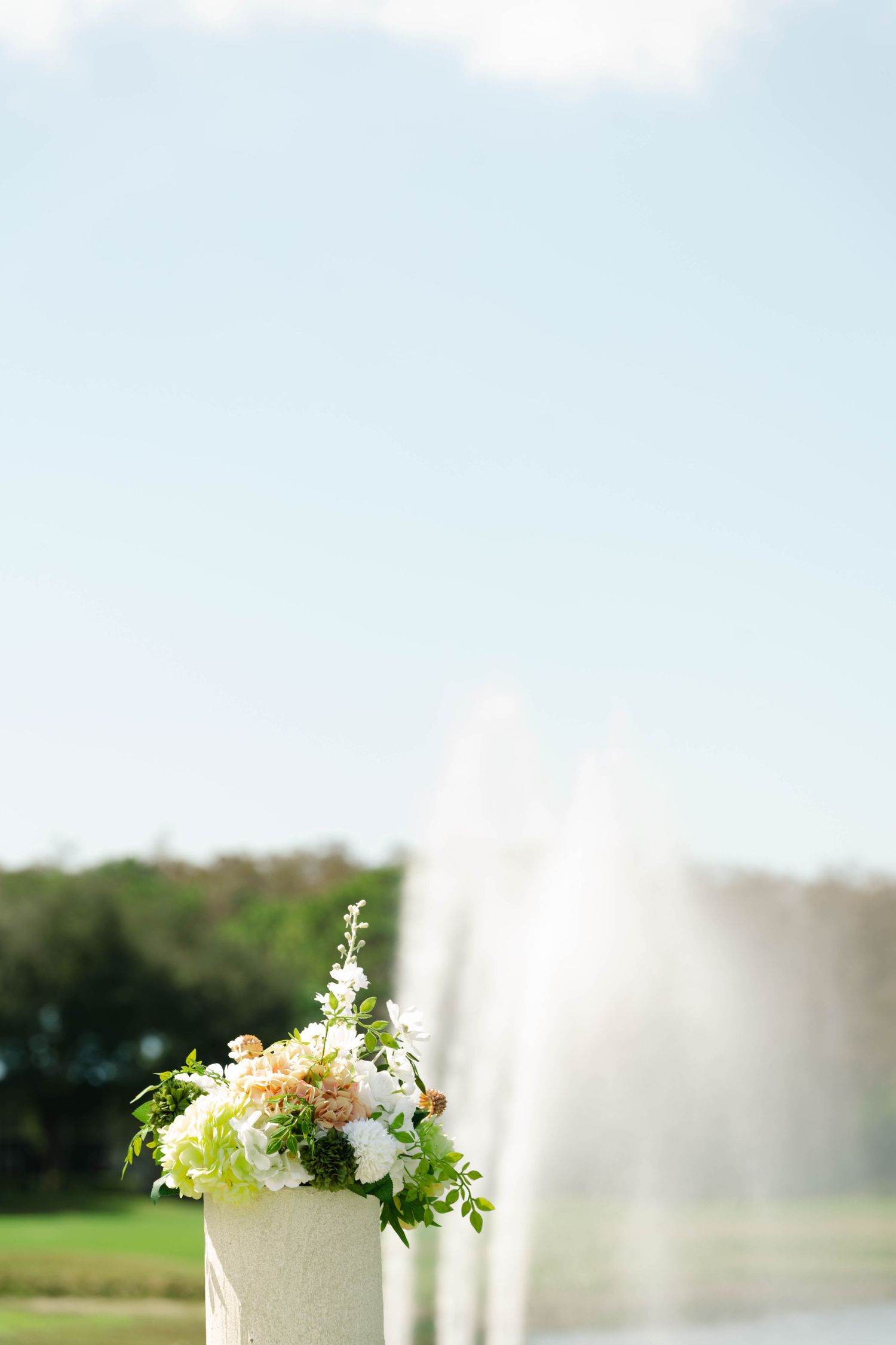 White and peach floral arrangement on a stone pedestal with a misty fountain and blue sky background.