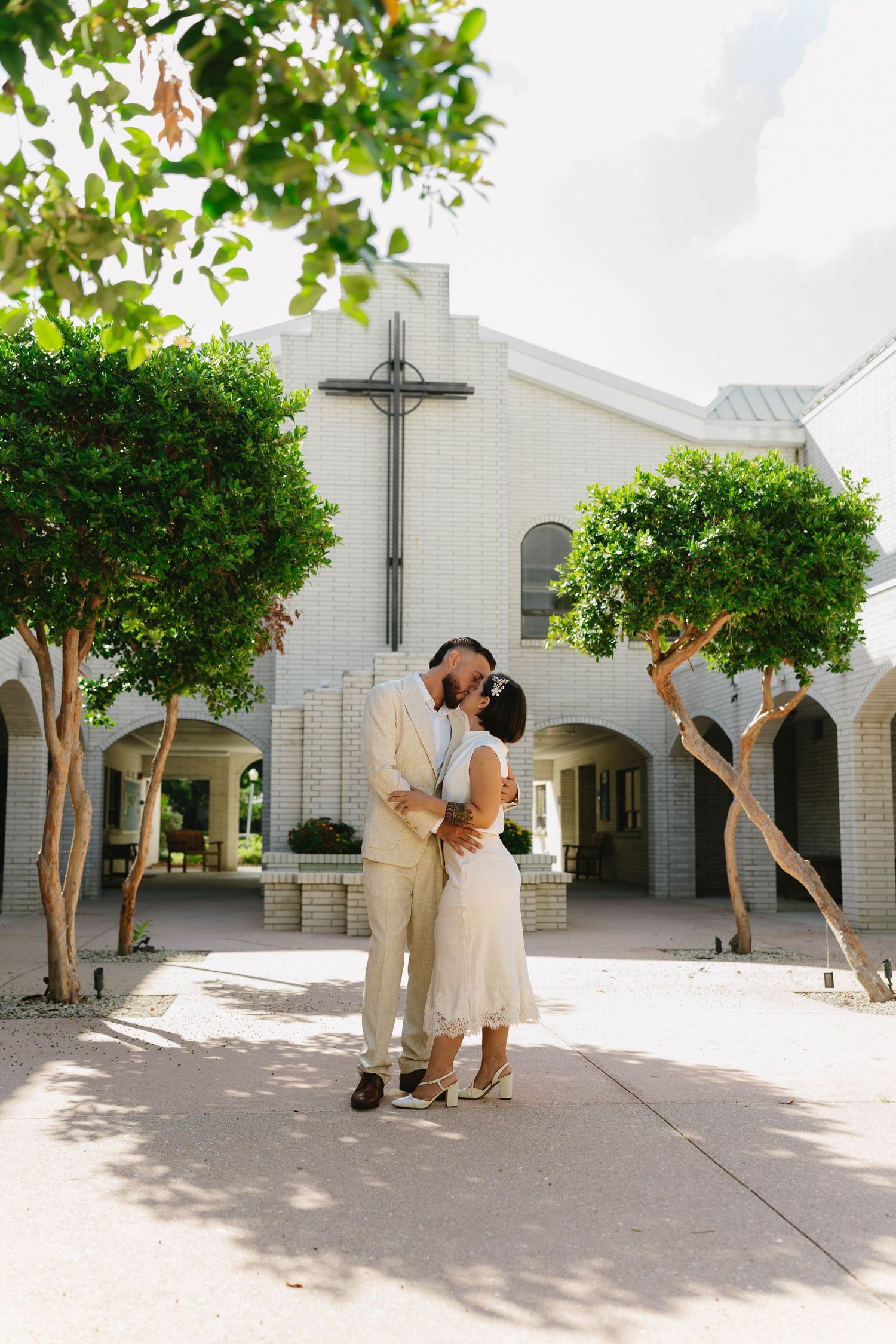 Couple shares kiss outside white brick church with cross, surrounded by manicured green trees on sunny day.