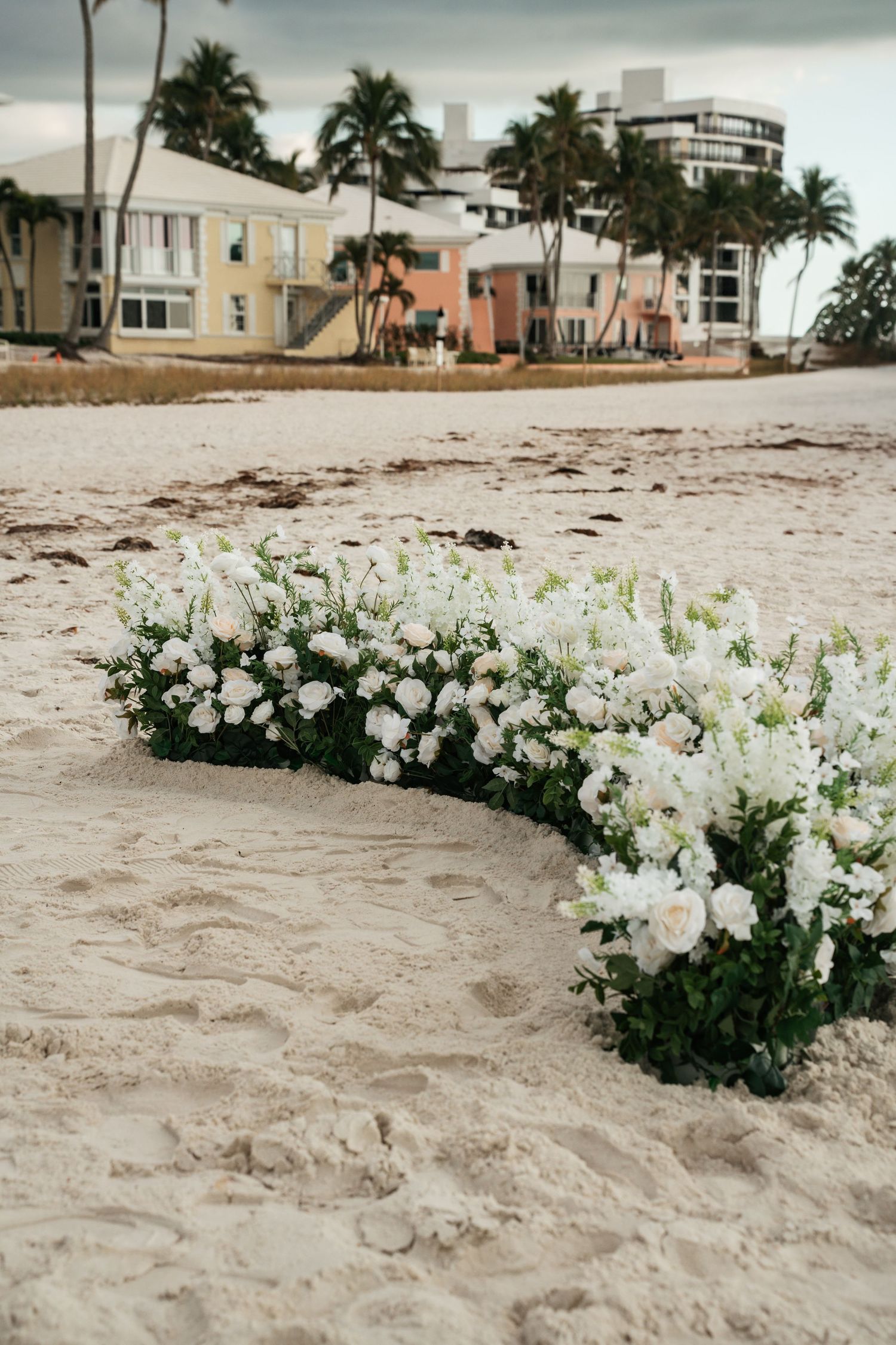 Curved arrangement of white roses and greenery decorating sandy beach wedding ceremony setup.