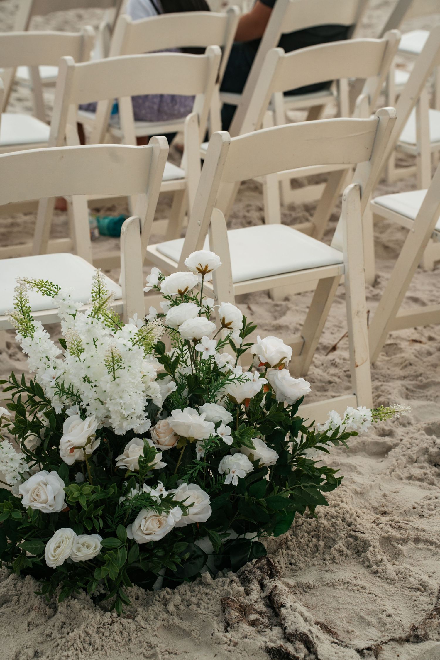 White roses and greenery floral arrangement placed in sand beside white chairs at beach wedding.