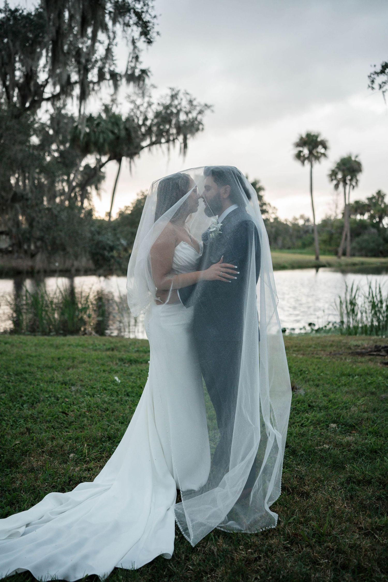 Bride and groom kiss under flowing veil beside serene lake with Spanish moss trees at dusk.