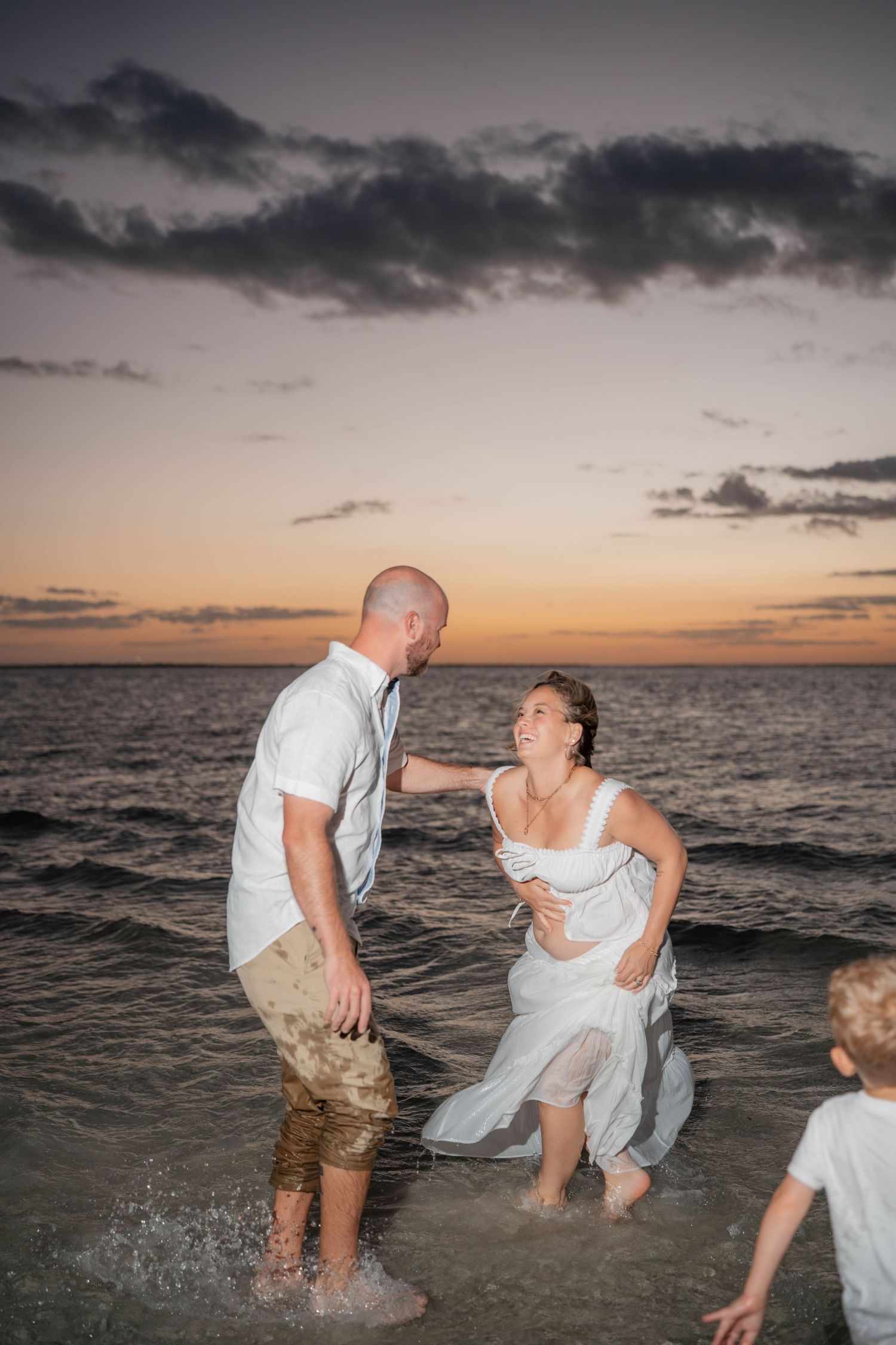 Couple in white outfits playfully splash in ocean waves at colorful sunset with child nearby.