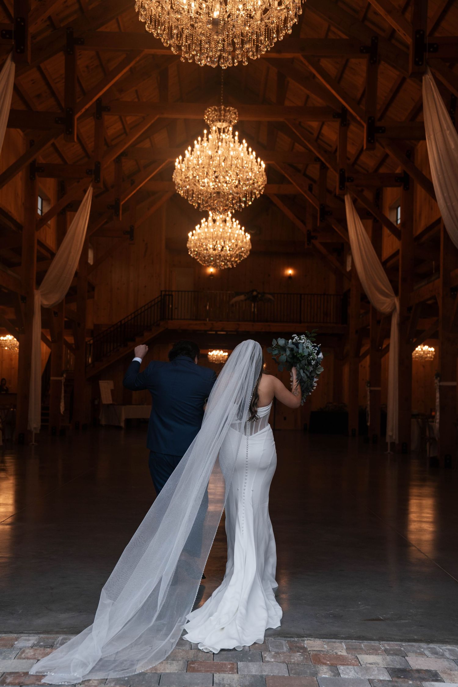 Bride and groom walk hand in hand inside elegant barn venue beneath cascading crystal chandeliers.