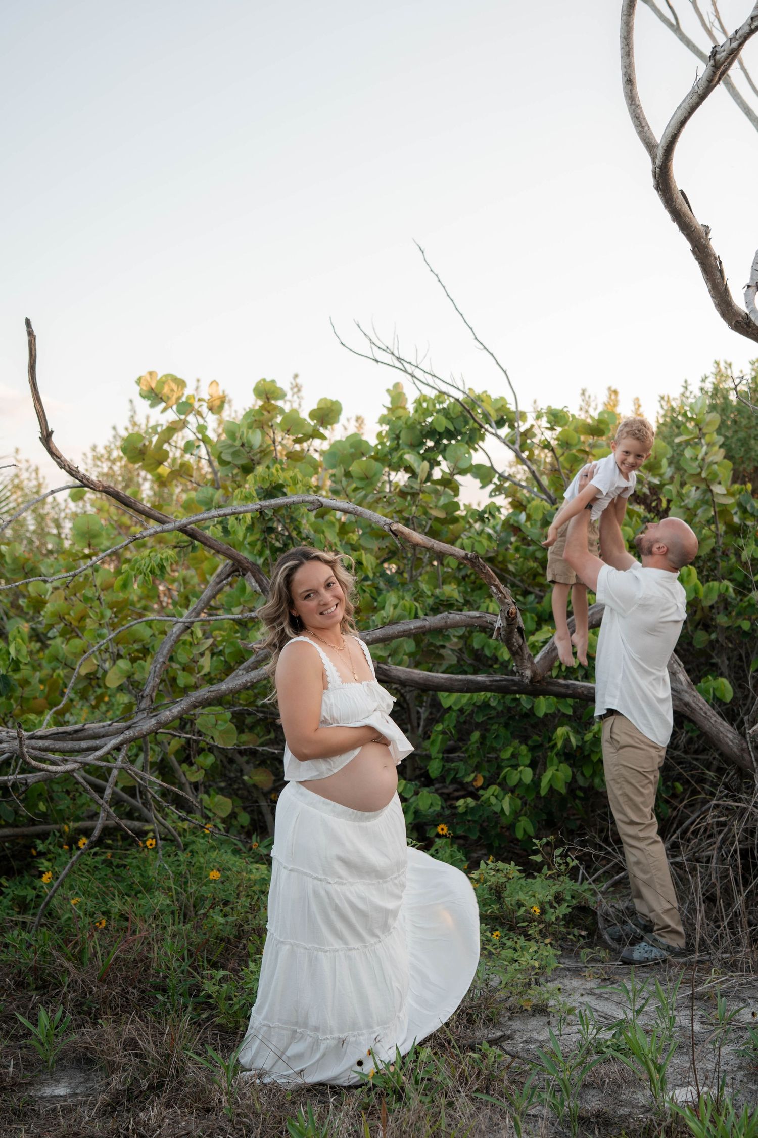 Pregnant woman in white dress poses while man lifts toddler over fallen trees on a sunny beach.