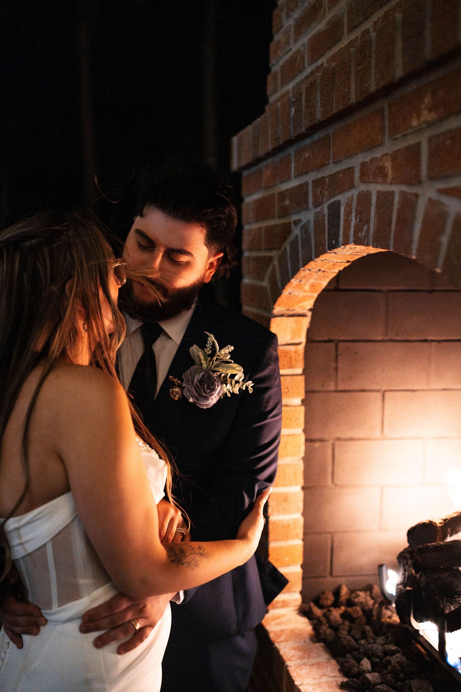 Bride and groom share intimate romantic moment by glowing brick fireplace.