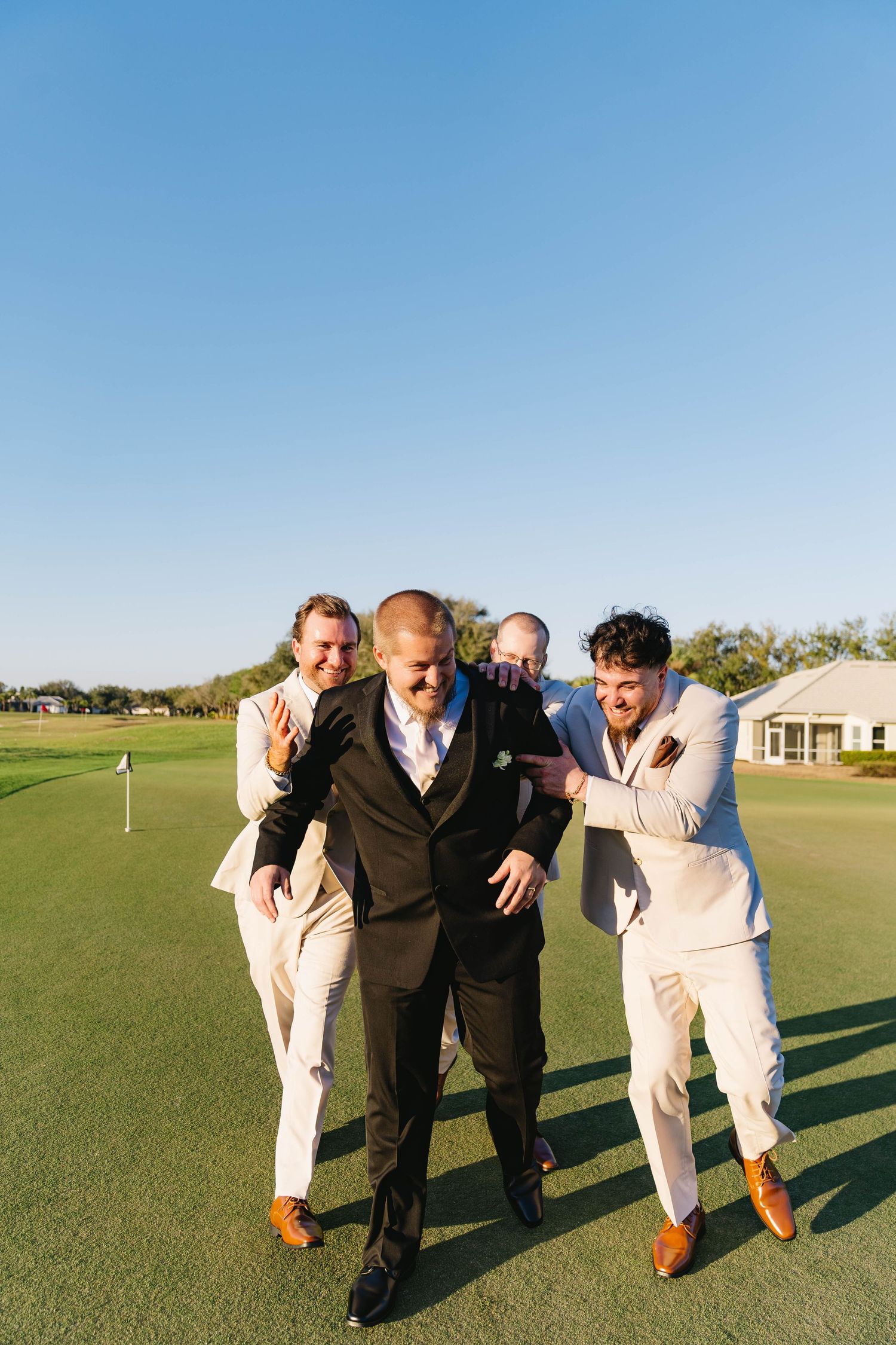 Groom in black suit walks with groomsmen in tan suits on sunny golf course green.