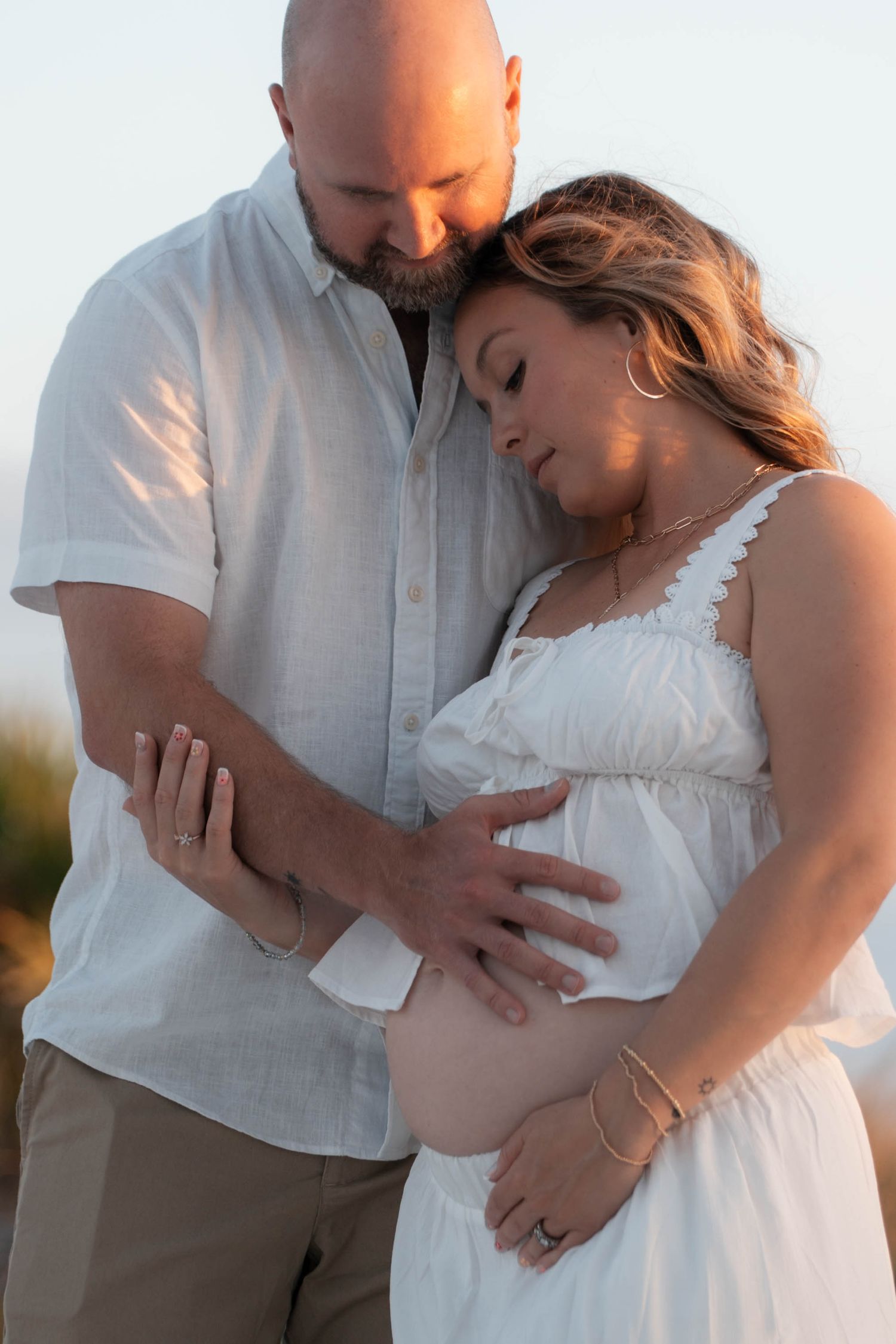 A couple tenderly cradles a pregnant belly outdoors at golden hour, both dressed in white and khaki.