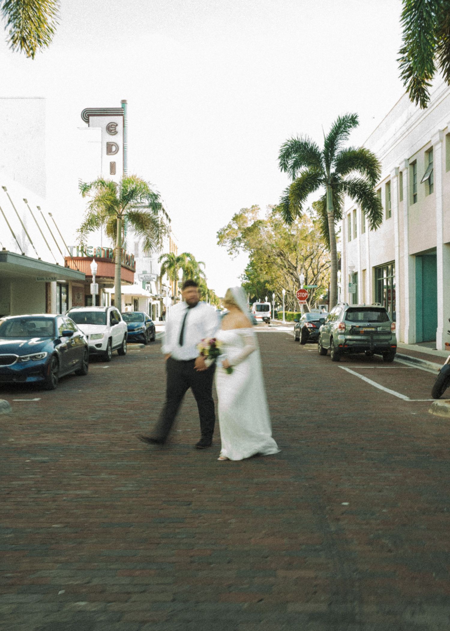 A bride and groom walk hand-in-hand across a brick street lined with palm trees in a sunny downtown setting.