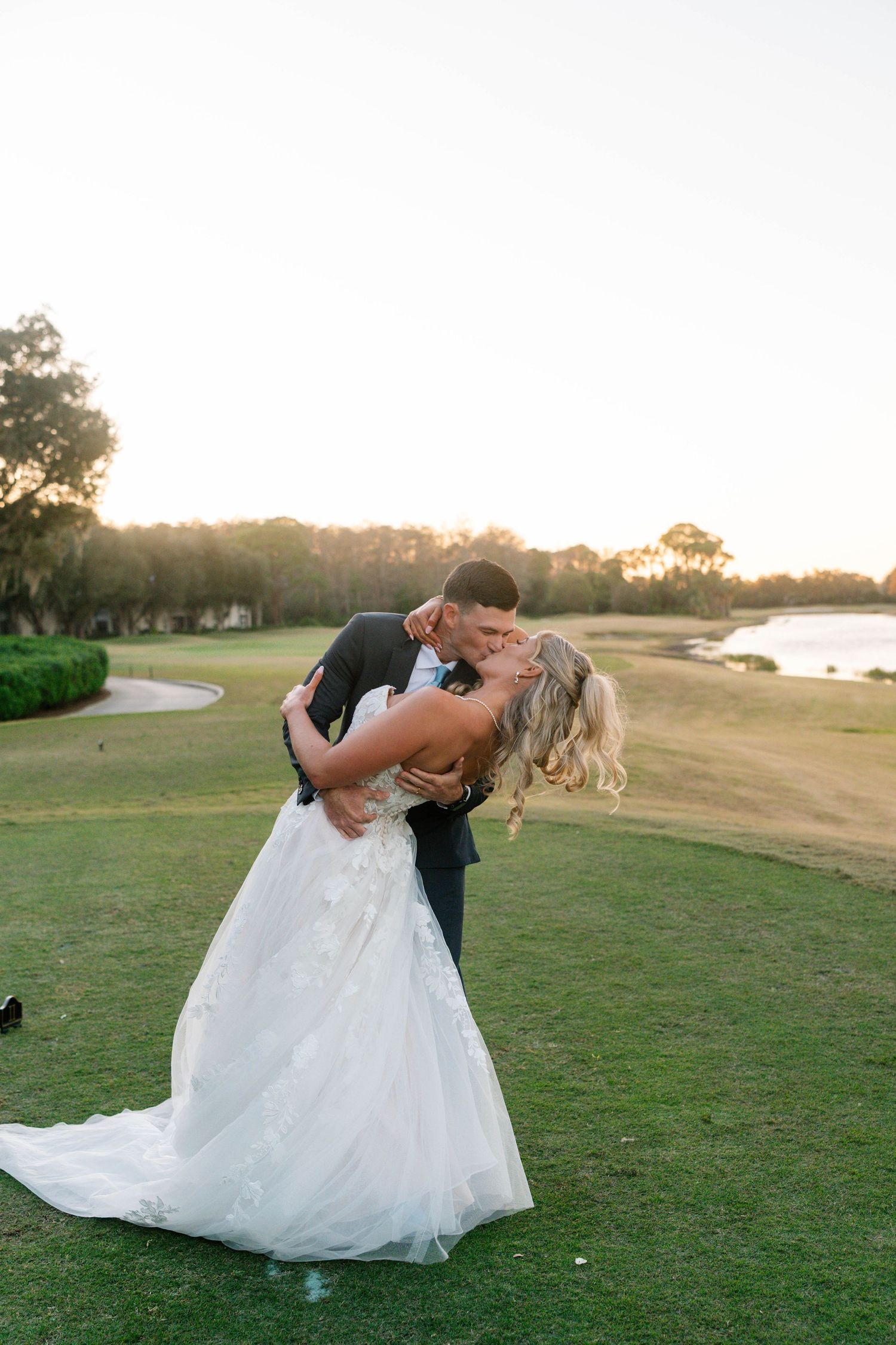 Groom dips bride for a romantic kiss on a golf course lawn at sunset with water in background.