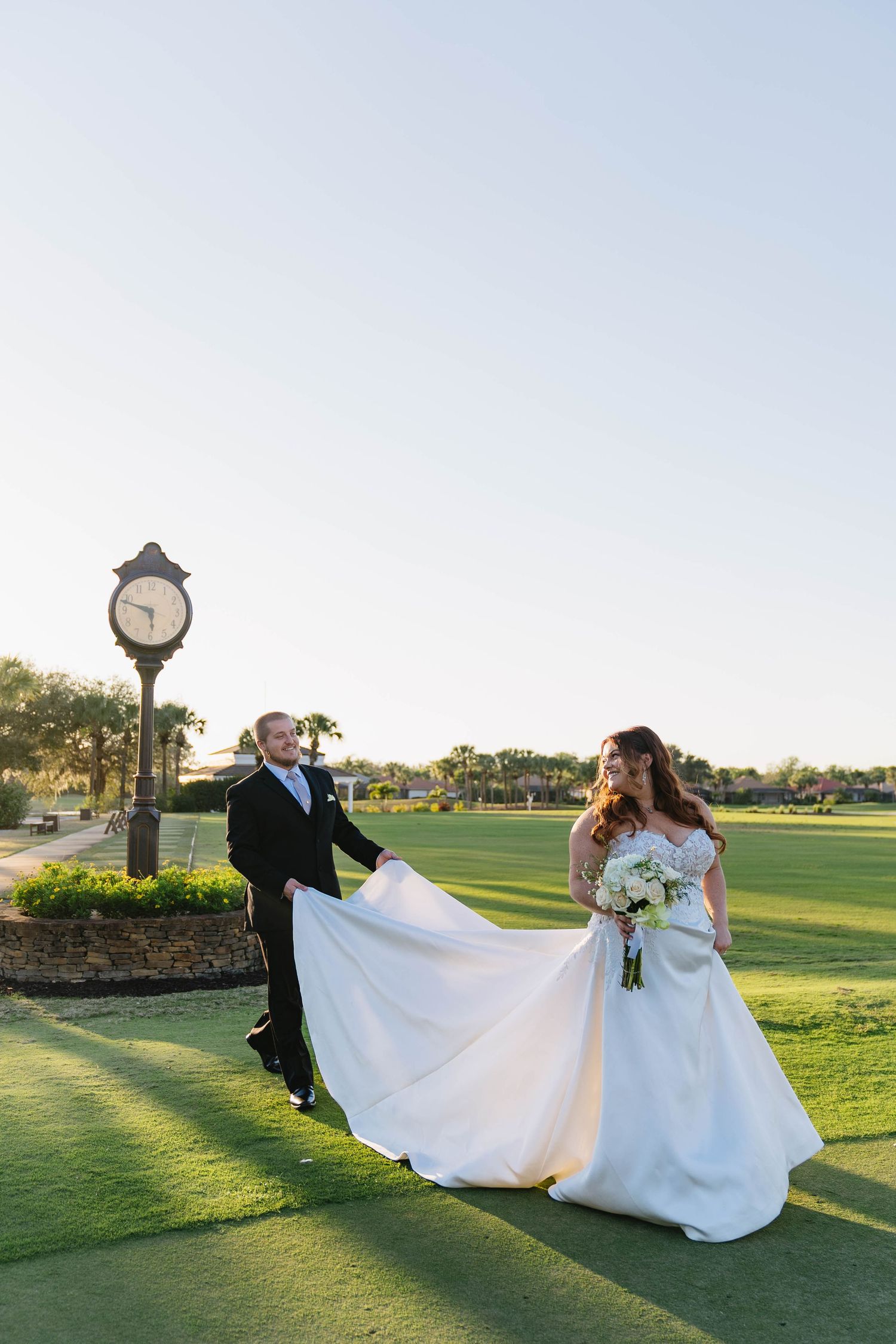 Bride and groom pose near vintage clock on golf course as groom lifts wedding gown.