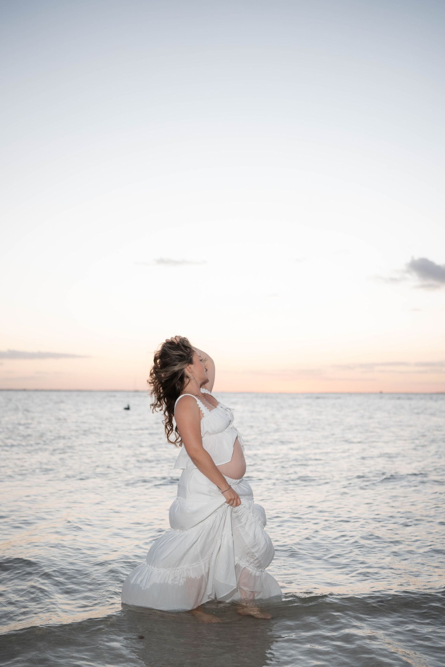 Pregnant woman in flowing white dress standing in ocean waves at sunset, joyfully embracing her baby bump.
