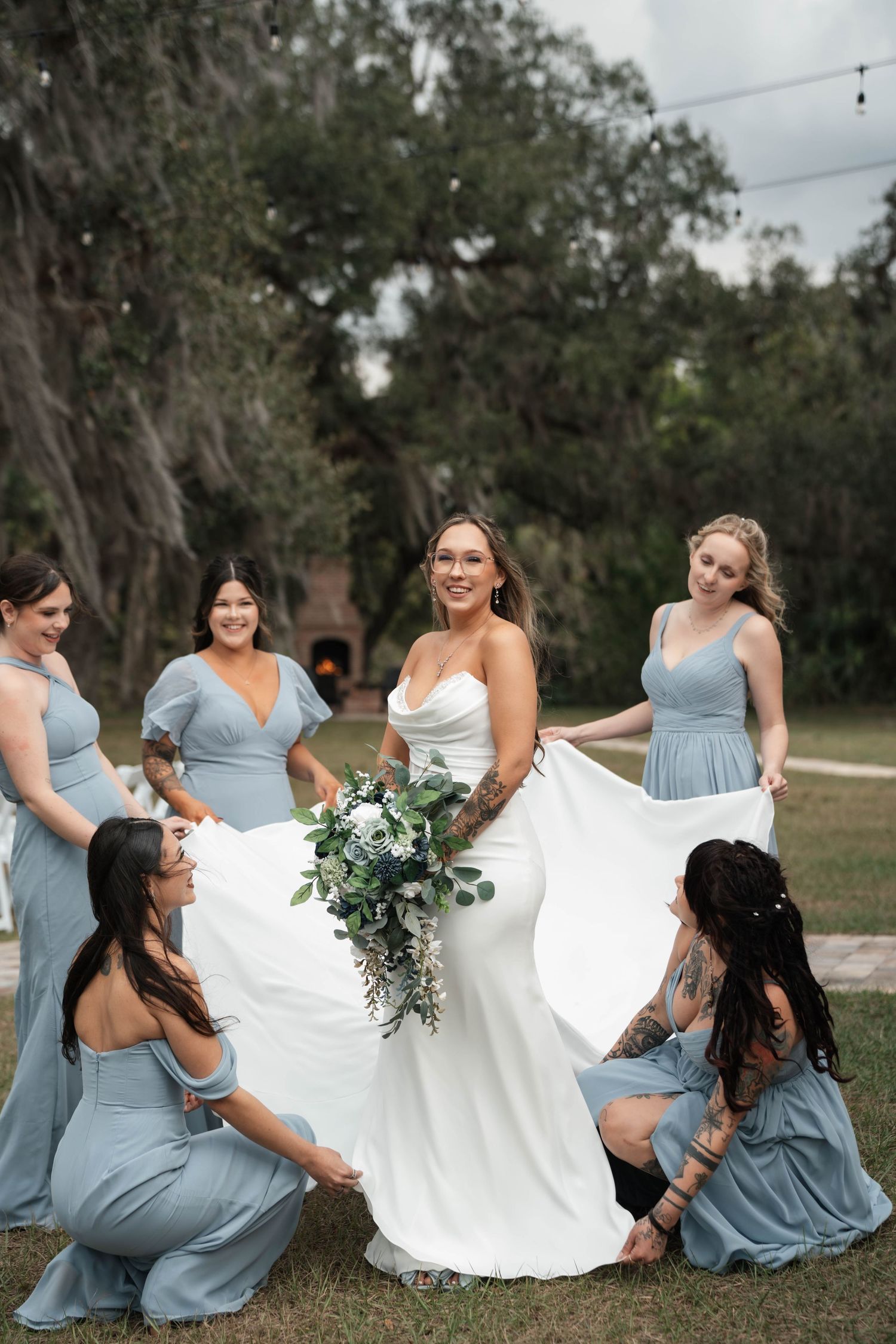 Bride smiles holding bouquet while bridesmaids in dusty blue dresses arrange her train.