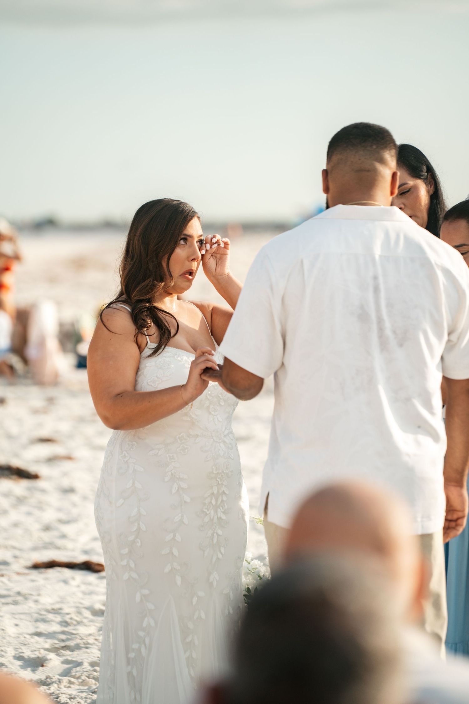 Emotional bride wiping tears during outdoor beach wedding ceremony at sunset.
