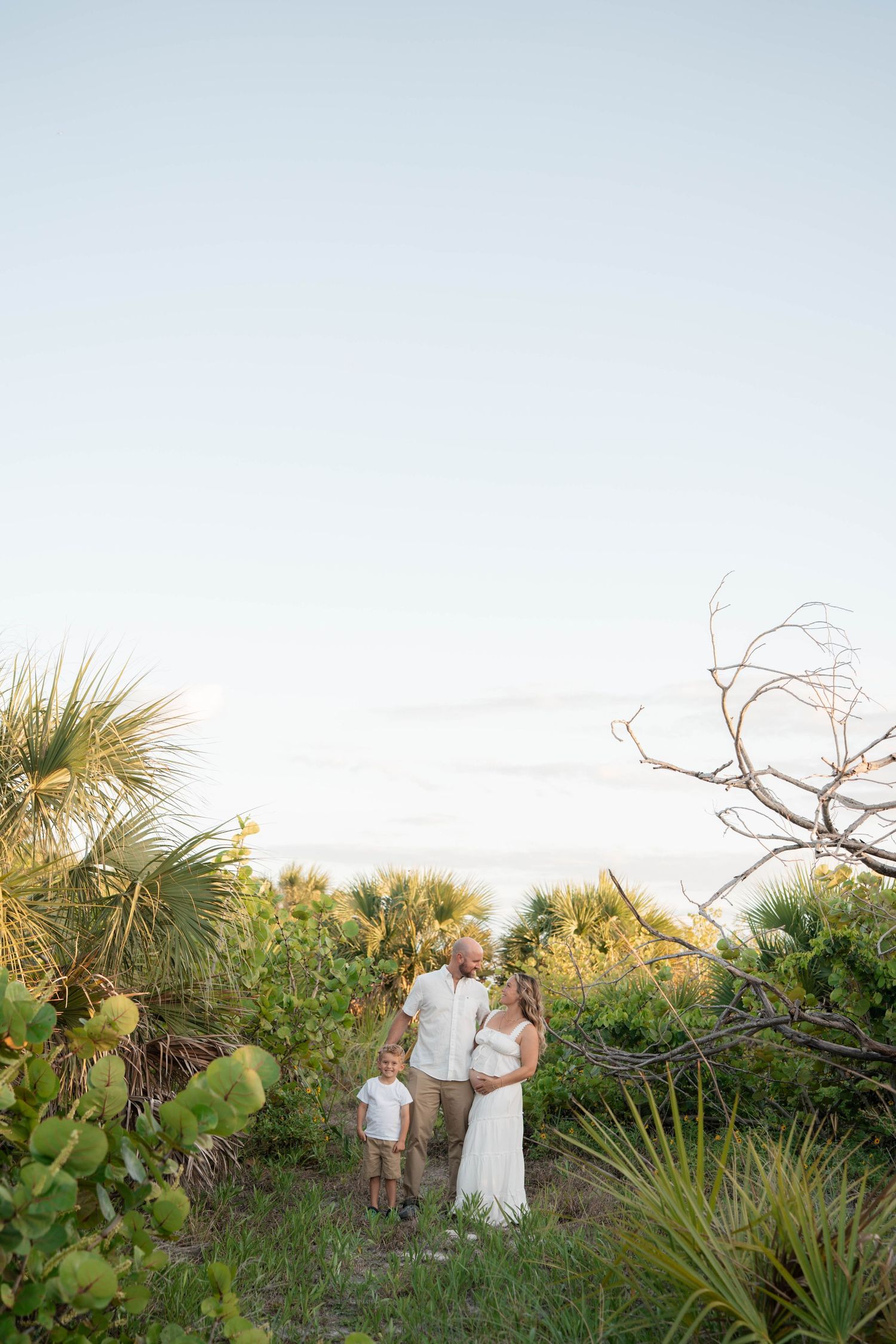 Family of three poses together among lush coastal vegetation at golden hour near the beach.