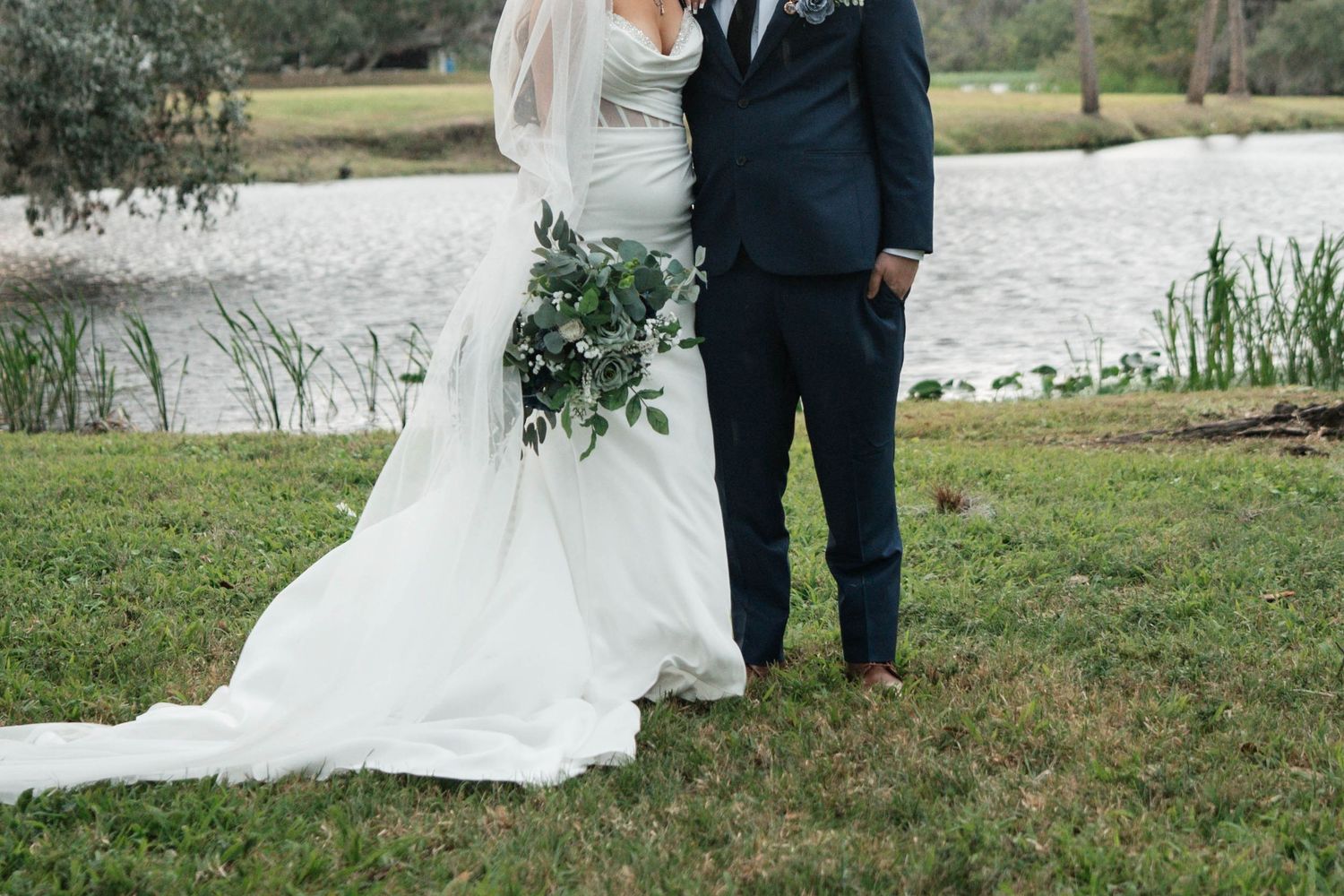 Wedding couple stands by waterfront showcasing brides cathedral train and lush greenery bouquet.