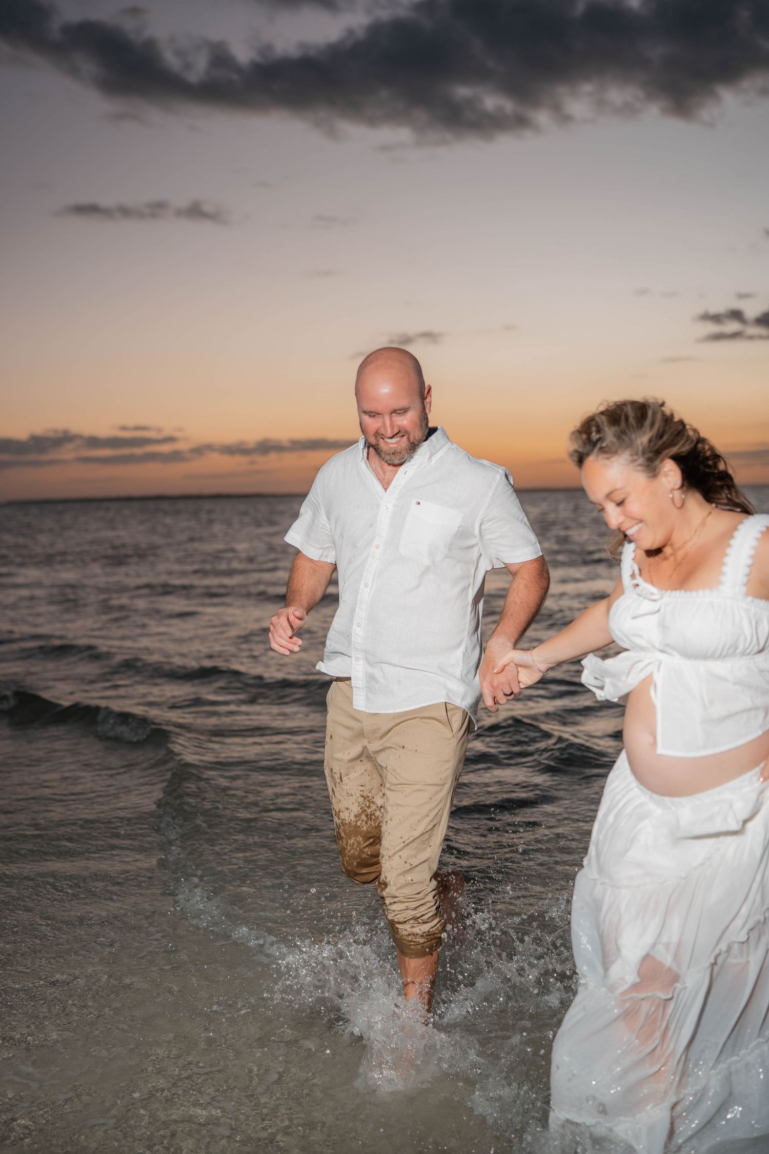 Couple in white outfits holding hands splashing in ocean waves at sunset beach.