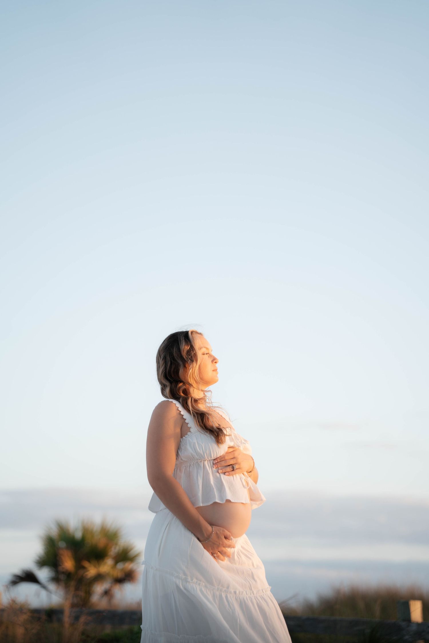 Pregnant woman in white dress cradles baby bump at golden hour with clear blue sky backdrop.