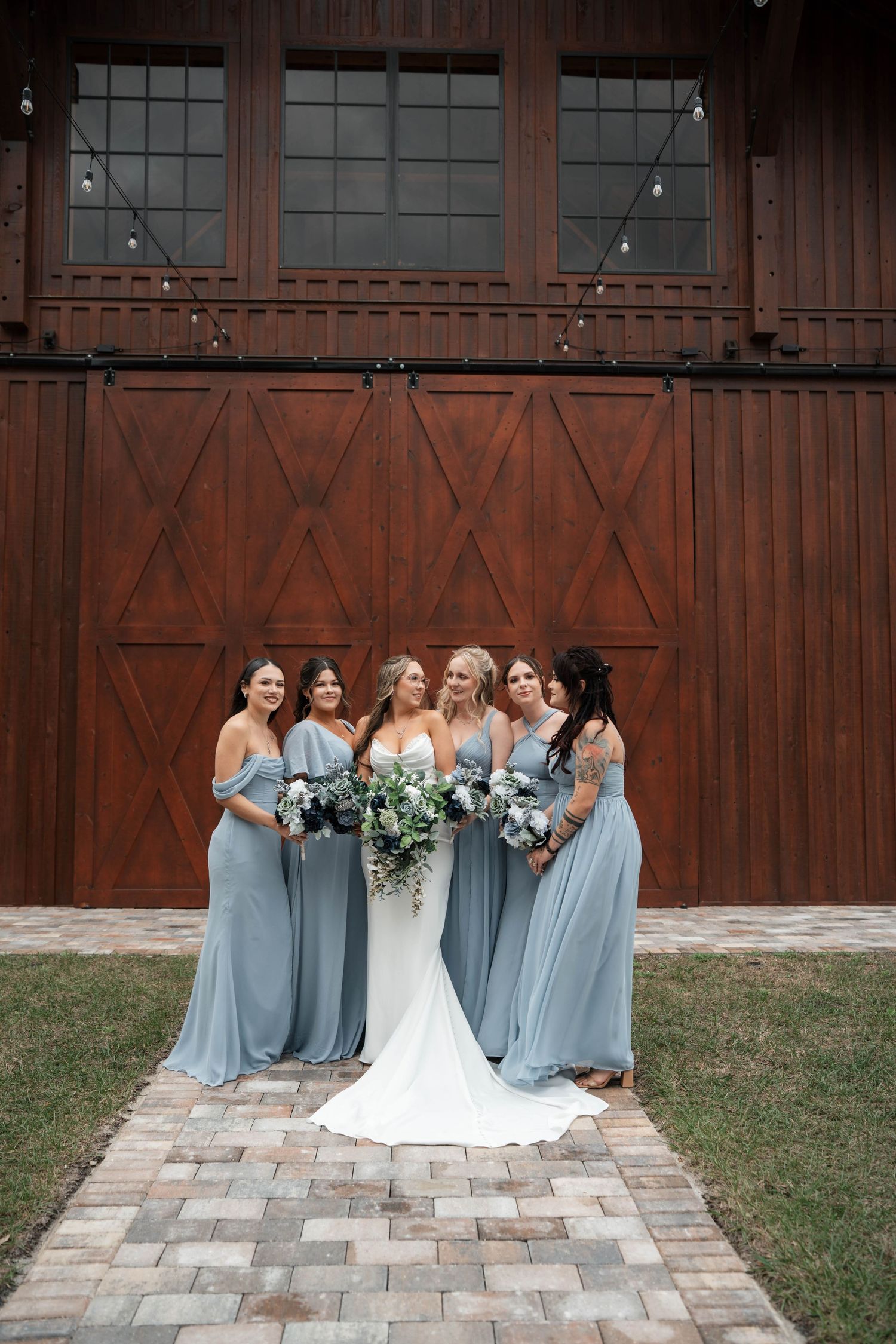 Bride with bridesmaids in dusty blue dresses posing in front of rustic barn doors.