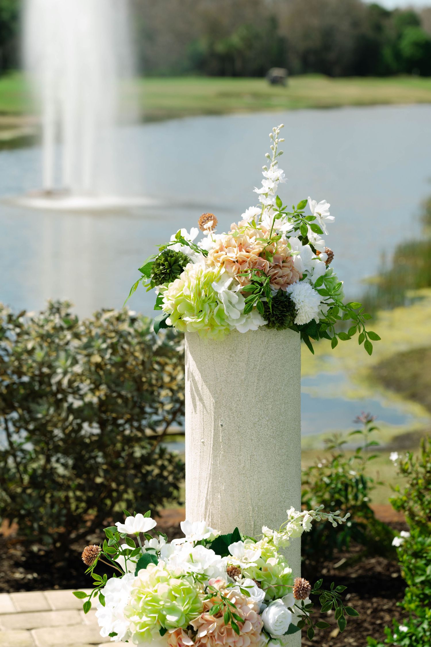 White and peach floral arrangement on a stone pedestal with a serene pond and fountain backdrop.