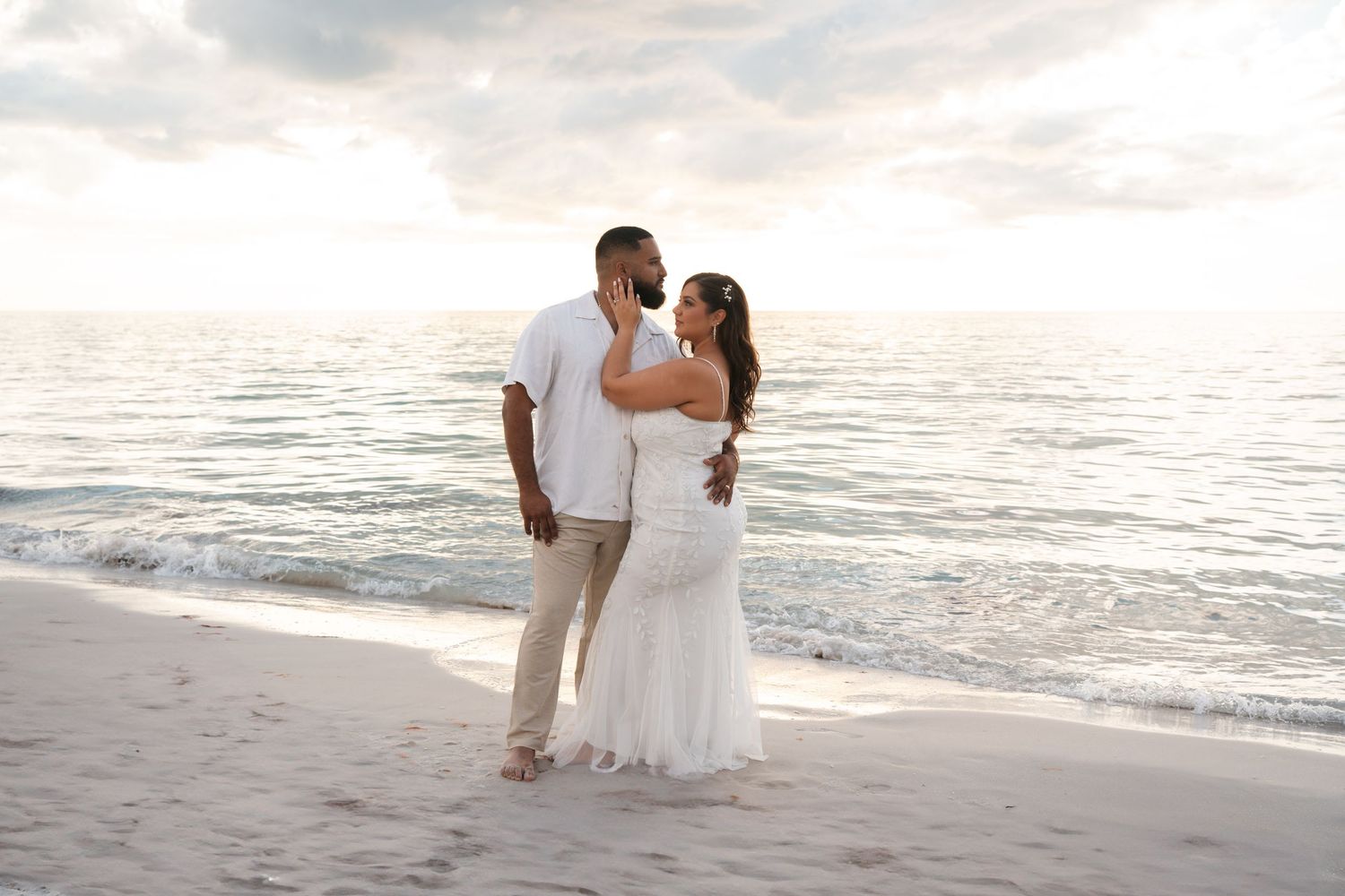 Newlywed couple embracing romantically on sandy beach at golden sunset by ocean.