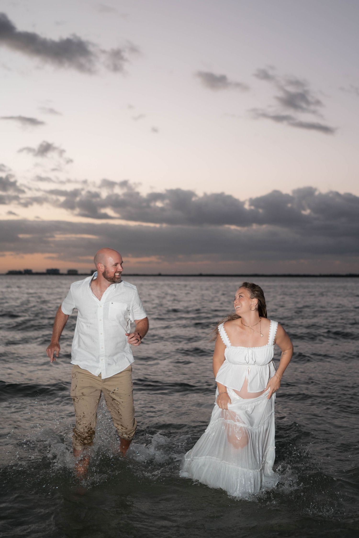 Couple in white outfits splashing through ocean waves at sunset with dramatic cloudy sky backdrop.