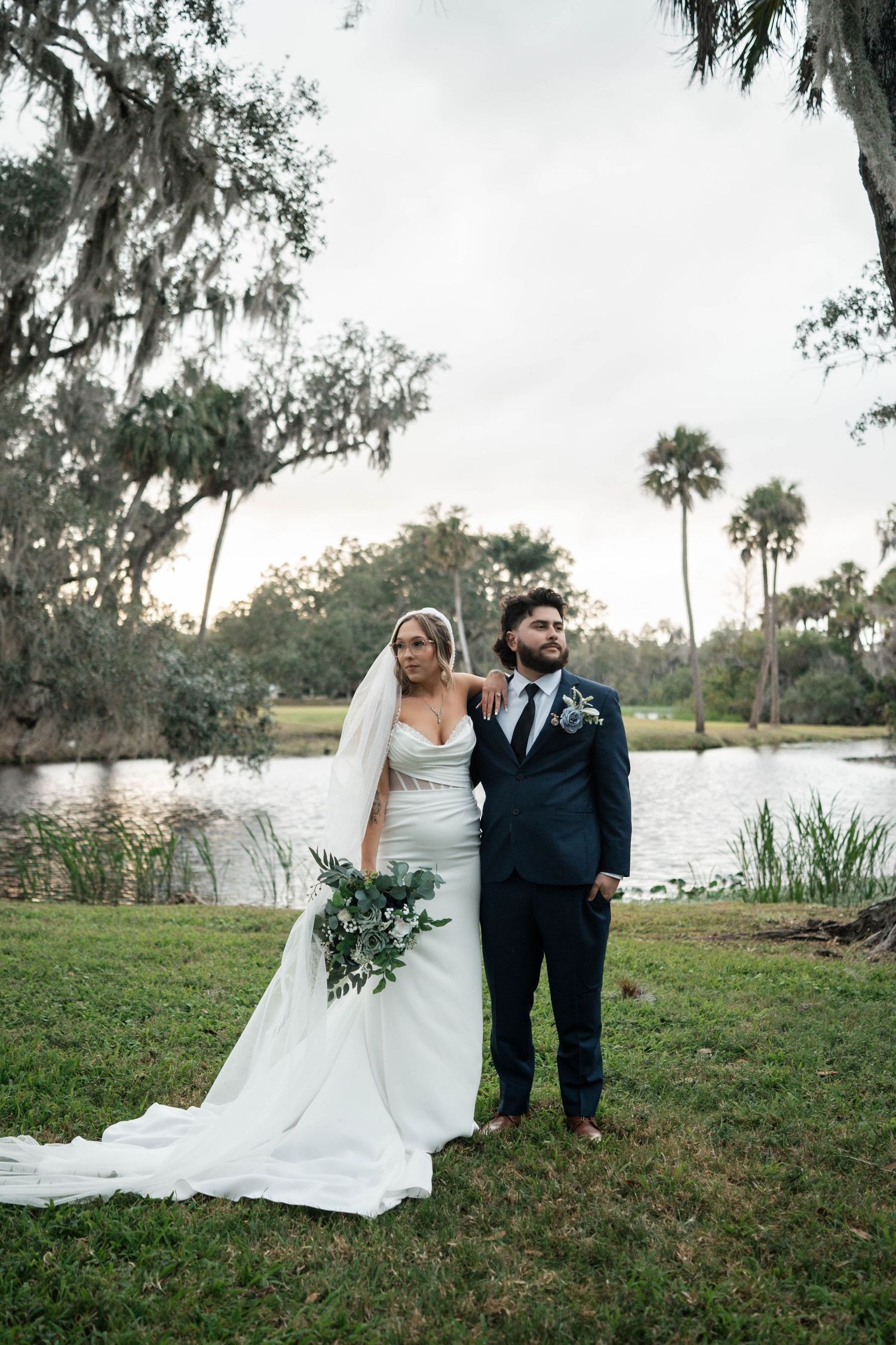 Elegant bride in fitted gown with long train and groom in navy suit pose by lakeside venue.
