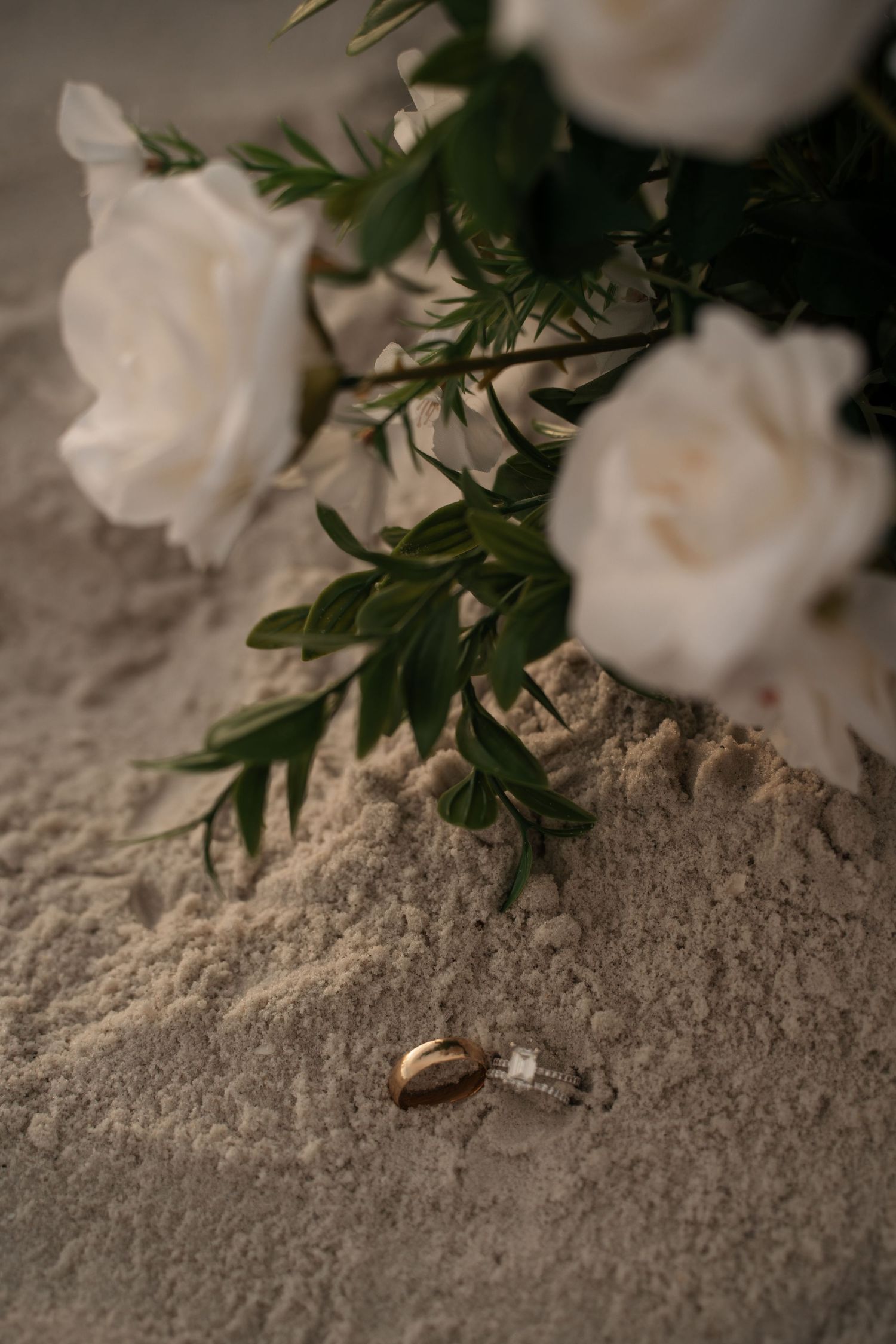 Two gold wedding rings resting in sand beside a bouquet of white roses and green foliage.