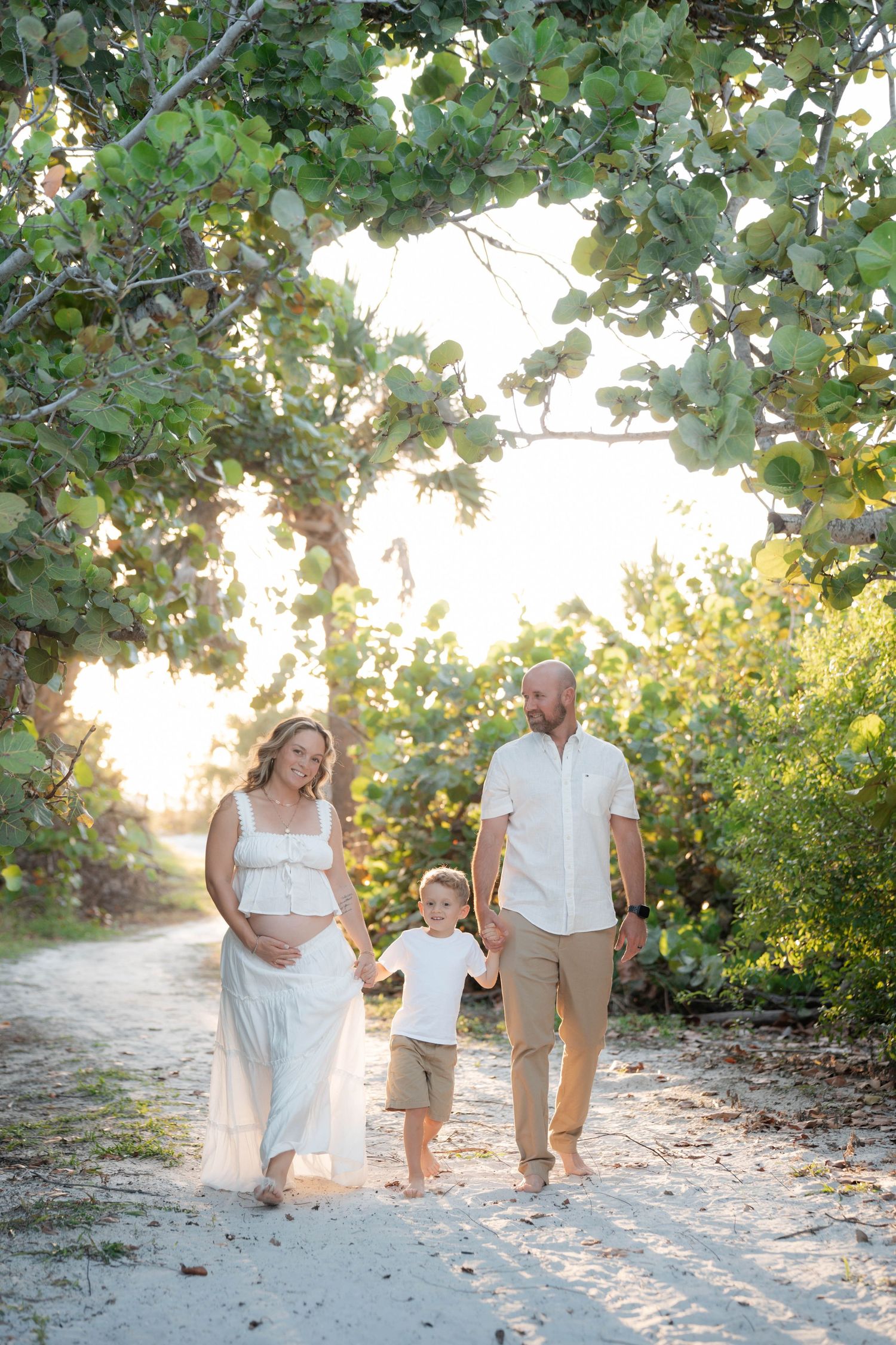 Pregnant woman in white dress walks with husband and young son on a sunlit sandy path framed by lush green trees.