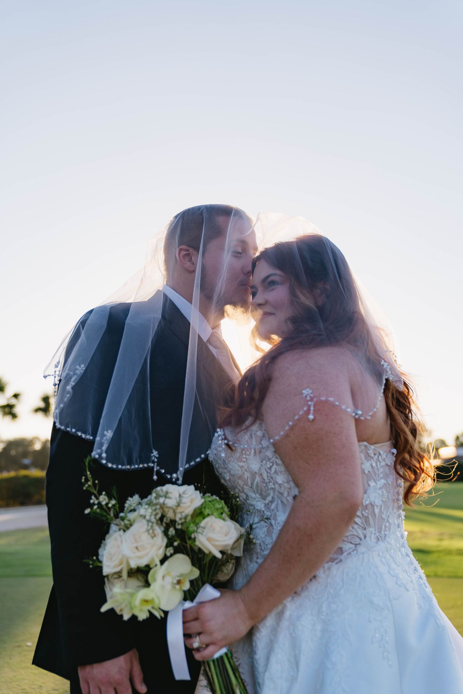 Bride and groom sharing a romantic kiss under a pearl-trimmed veil at golden hour on a golf course.